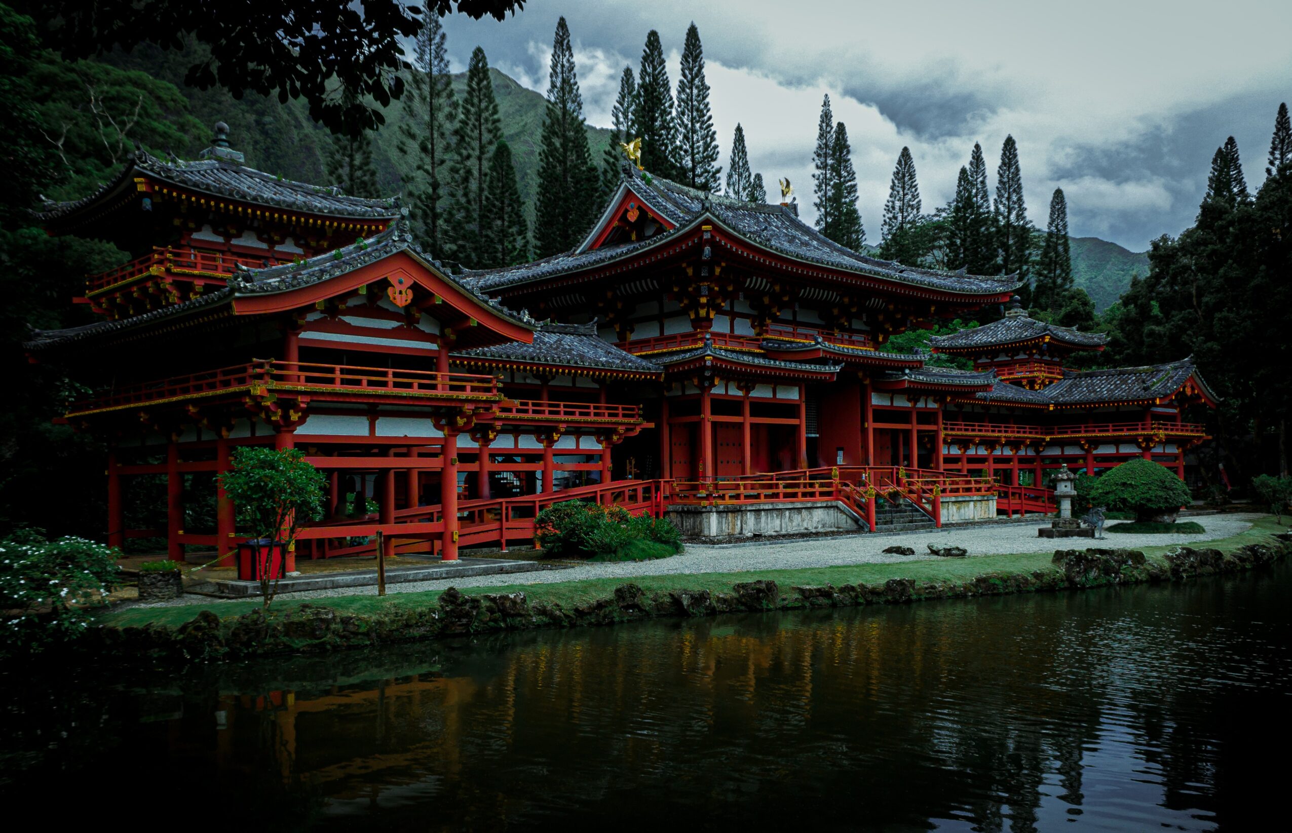Traditional Japanese building with red wooden structures, surrounded by trees and reflected in a body of water.