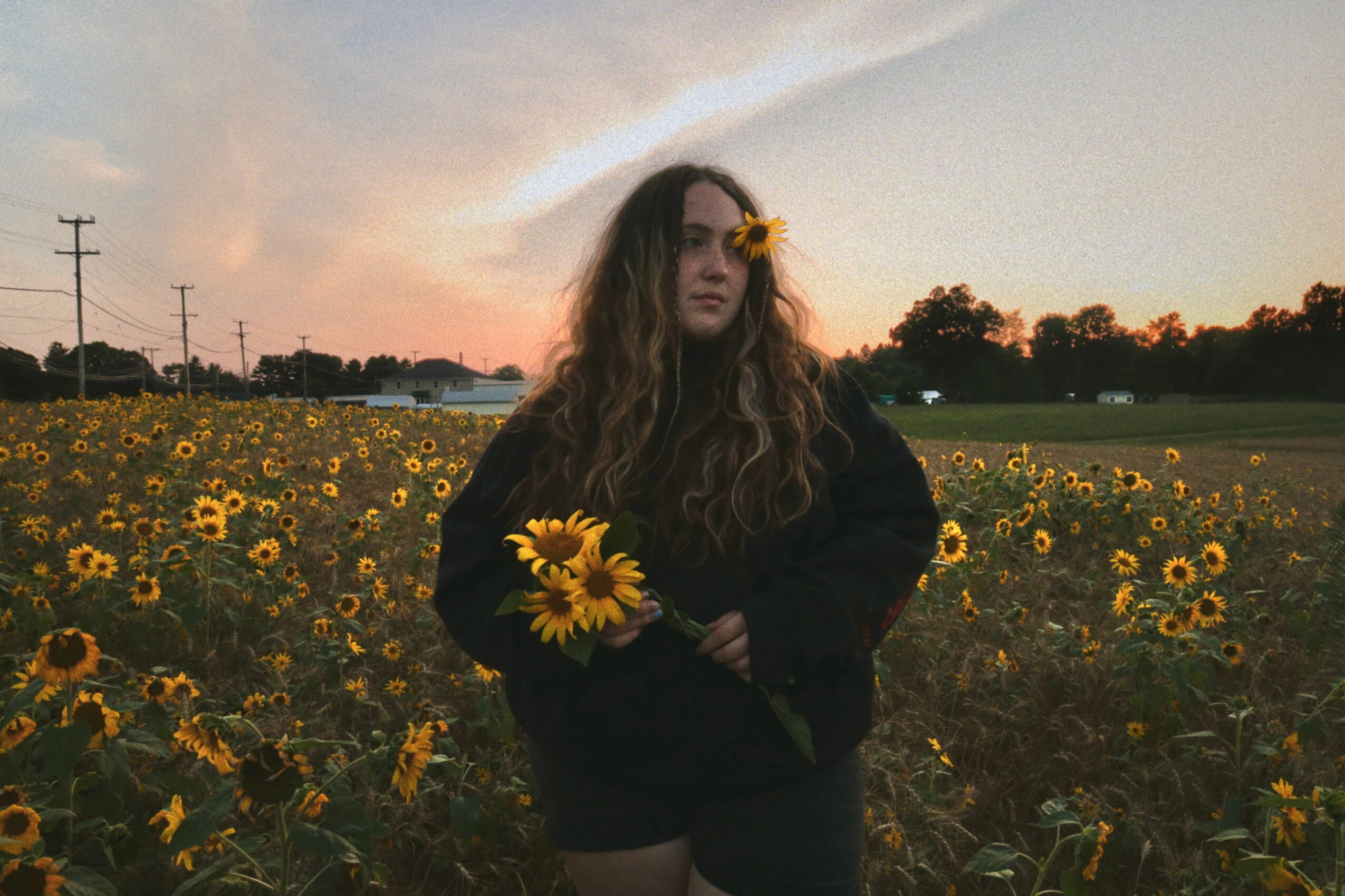 Young woman with long hair holding sunflowers in a sunflower field at sunset, with a flower in her hair.