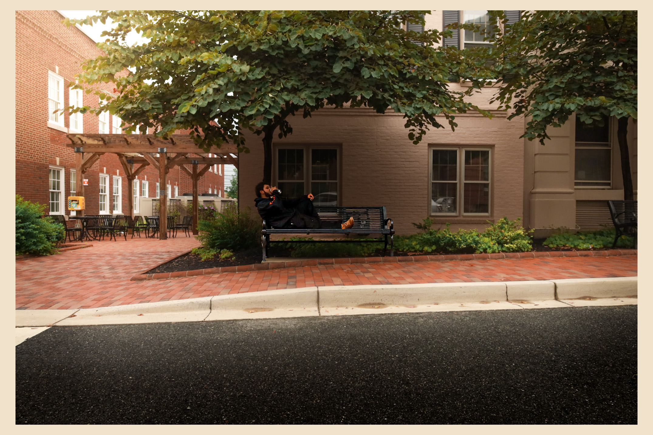 Two people sit on a bench under a tree in a courtyard with brick pavement and surrounding buildings.