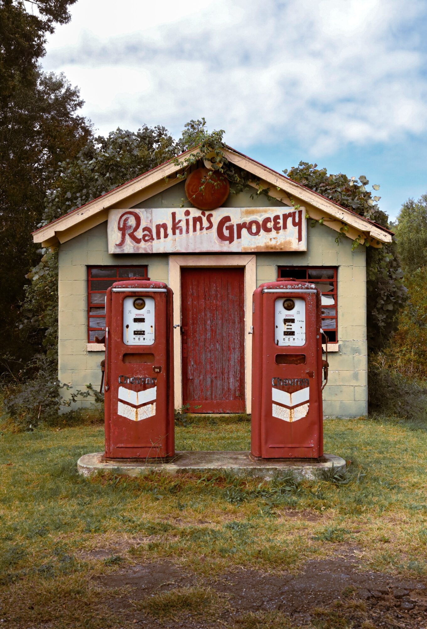 Small grocery store with two vintage gas pumps in front, sign reads 'Rankin's Grocery,' surrounded by grass and trees.