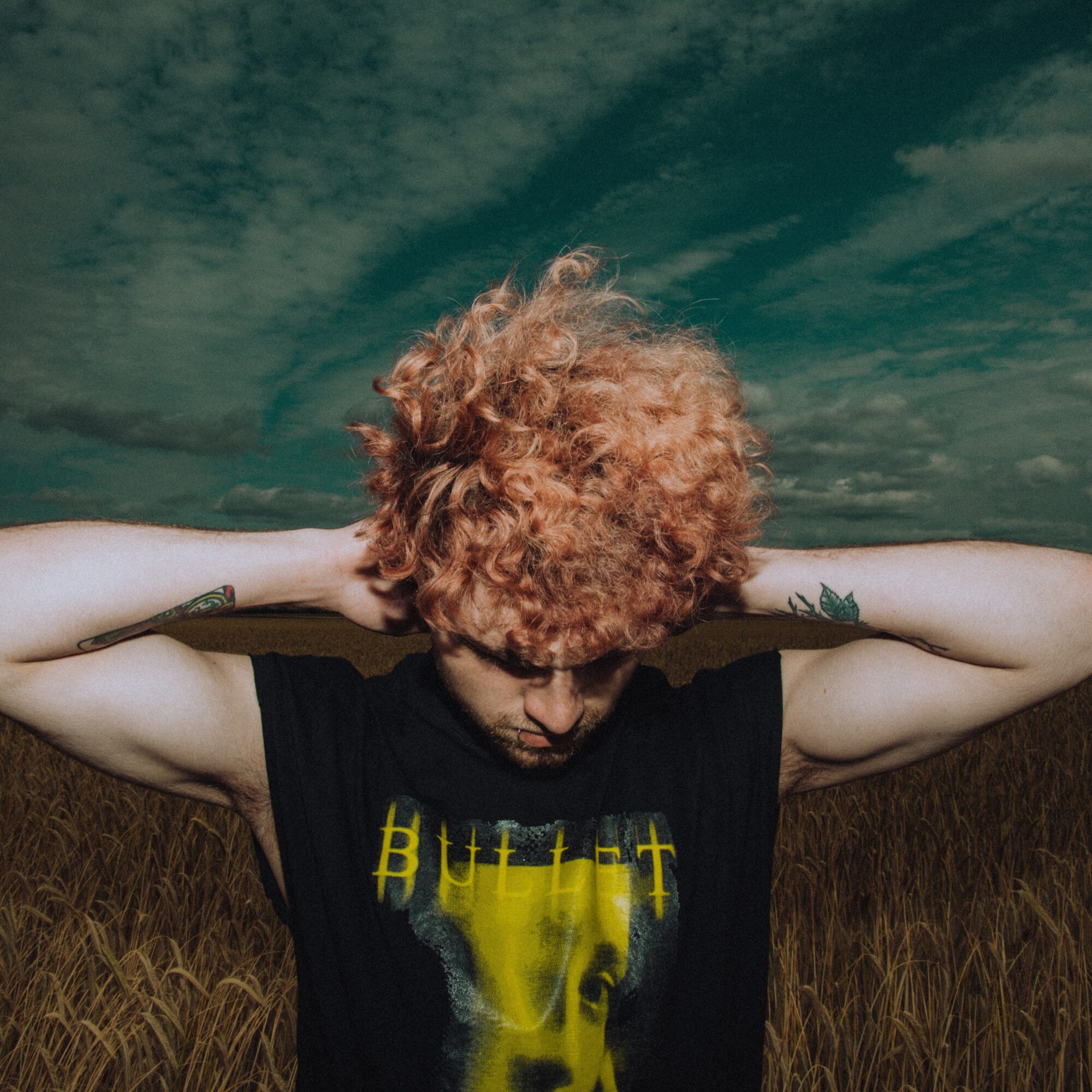 Person with curly red hair and tattoos, wearing a black T-shirt, standing outdoors with a cloudy sky background.