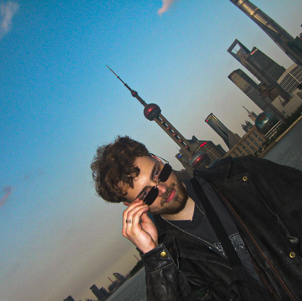 Man with curly hair and sunglasses adjusting his glasses, city skyline with tall buildings and a tower in the background.
