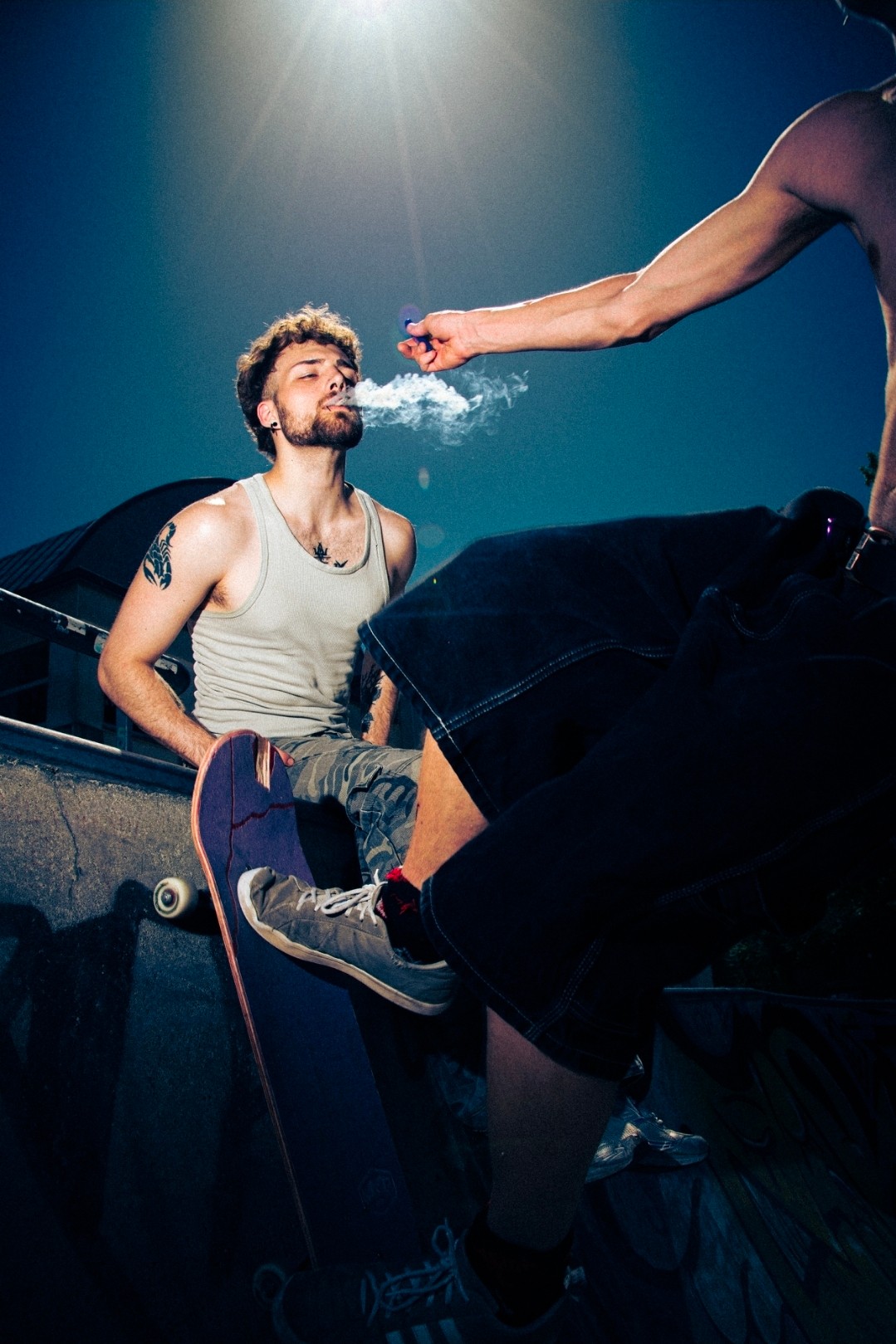 Person with a beard and tattoos sits on a stage, another person sprays water into their face, stage lighting above.