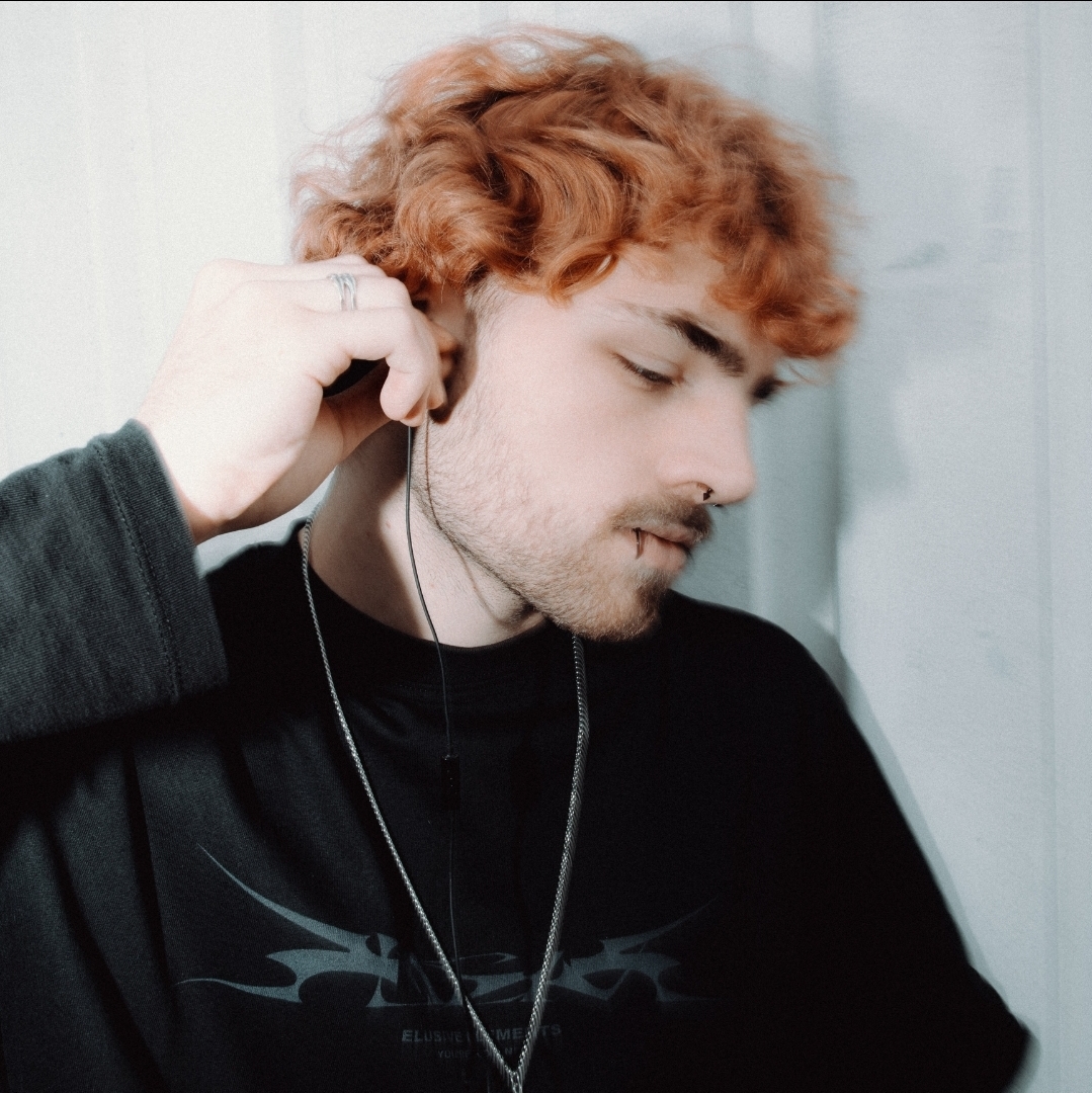 Young man with curly red hair and a beard, wearing headphones and a black shirt, looking down thoughtfully.