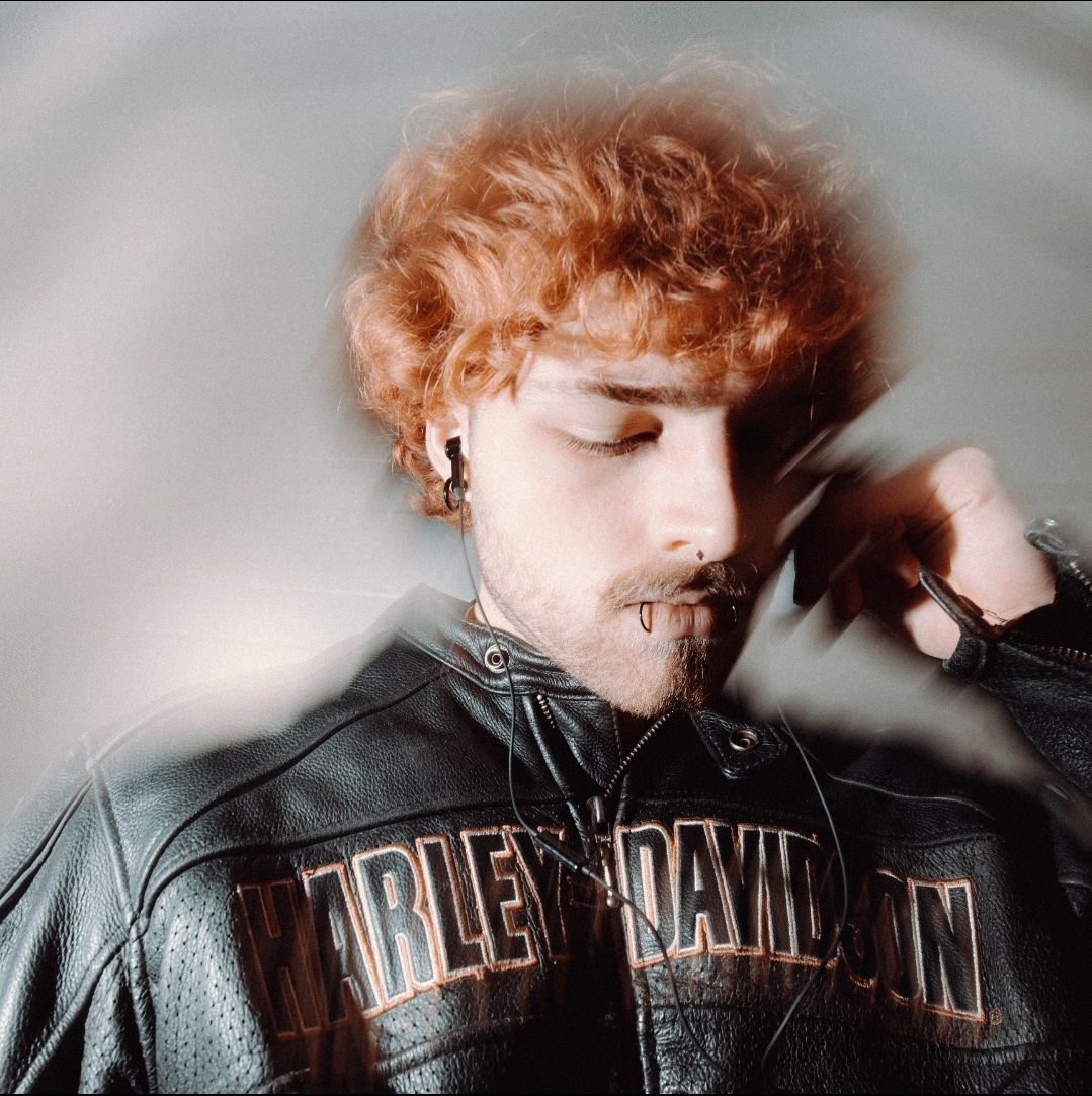Young man with curly orange hair wearing a black leather Harley Davidson jacket, eyes closed, touching his face.