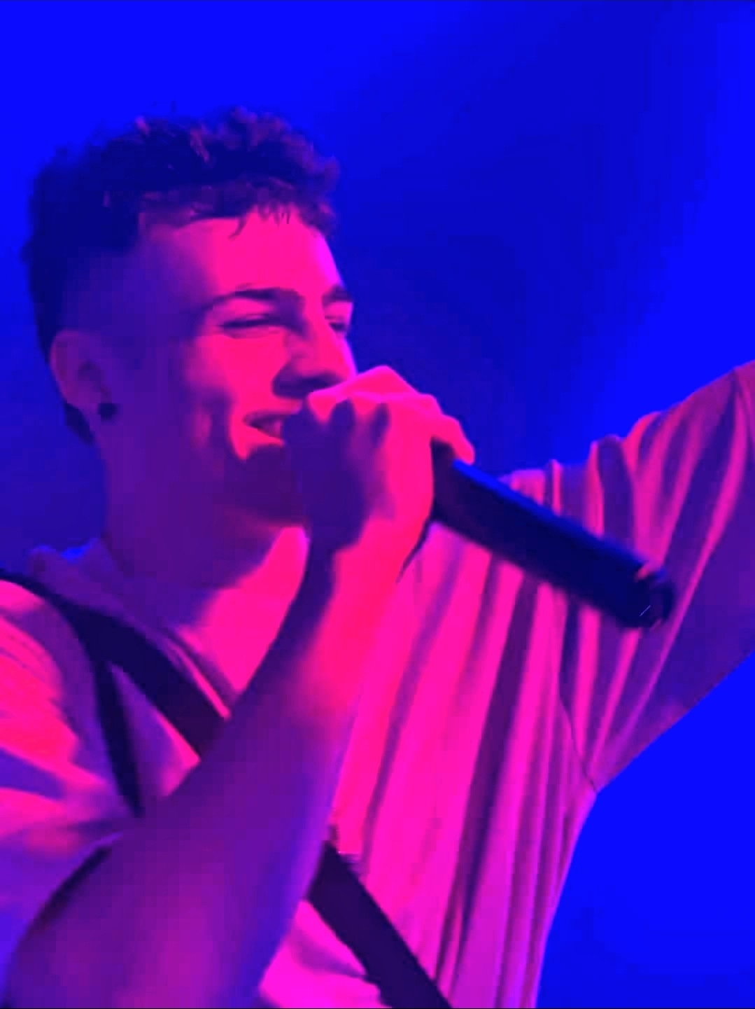 Person smiling and holding a microphone on stage with blue and pink lighting.
