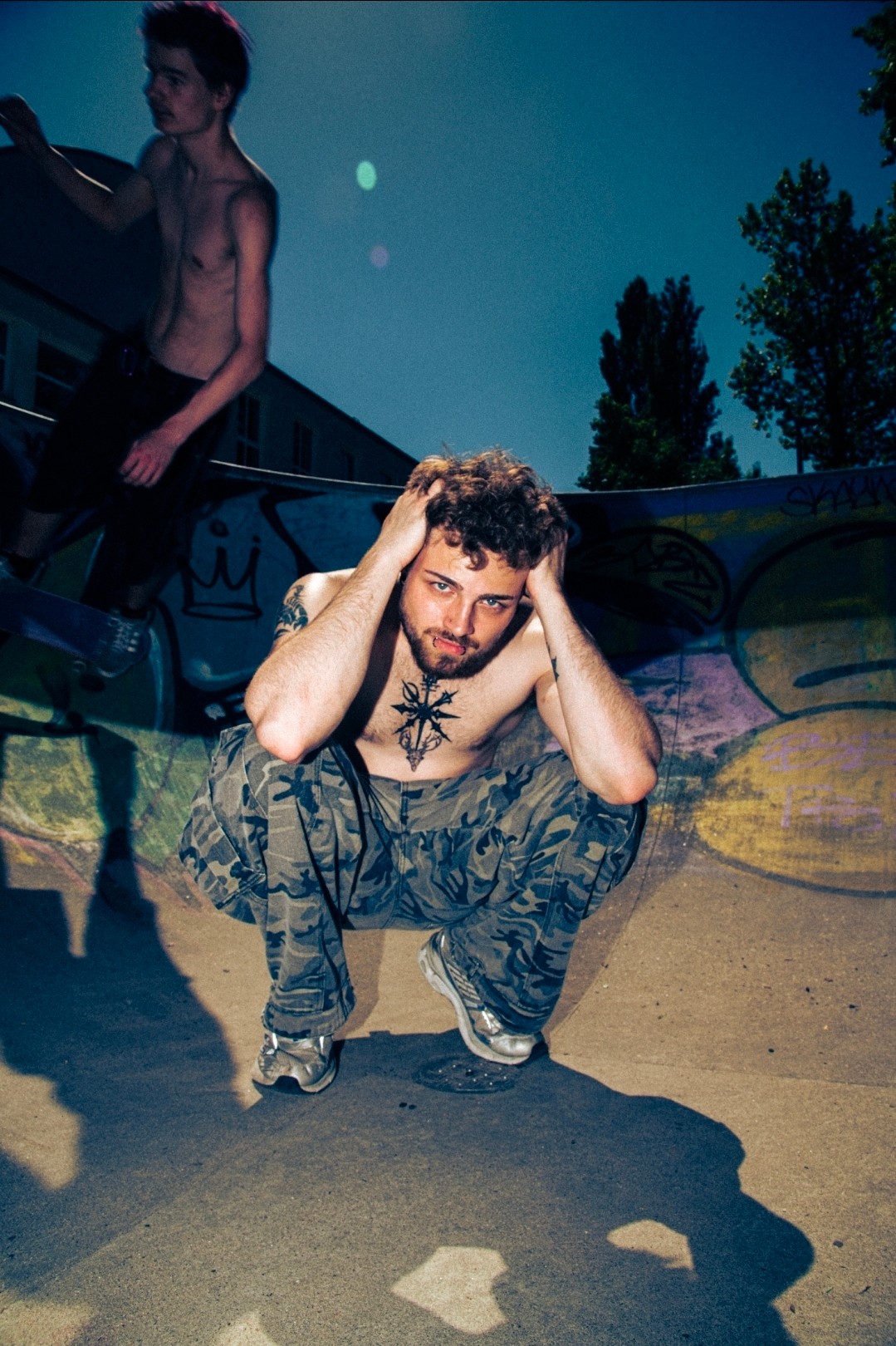 A man crouches with hands on head, wearing camouflage pants and a star necklace, at a skate park during daytime.
