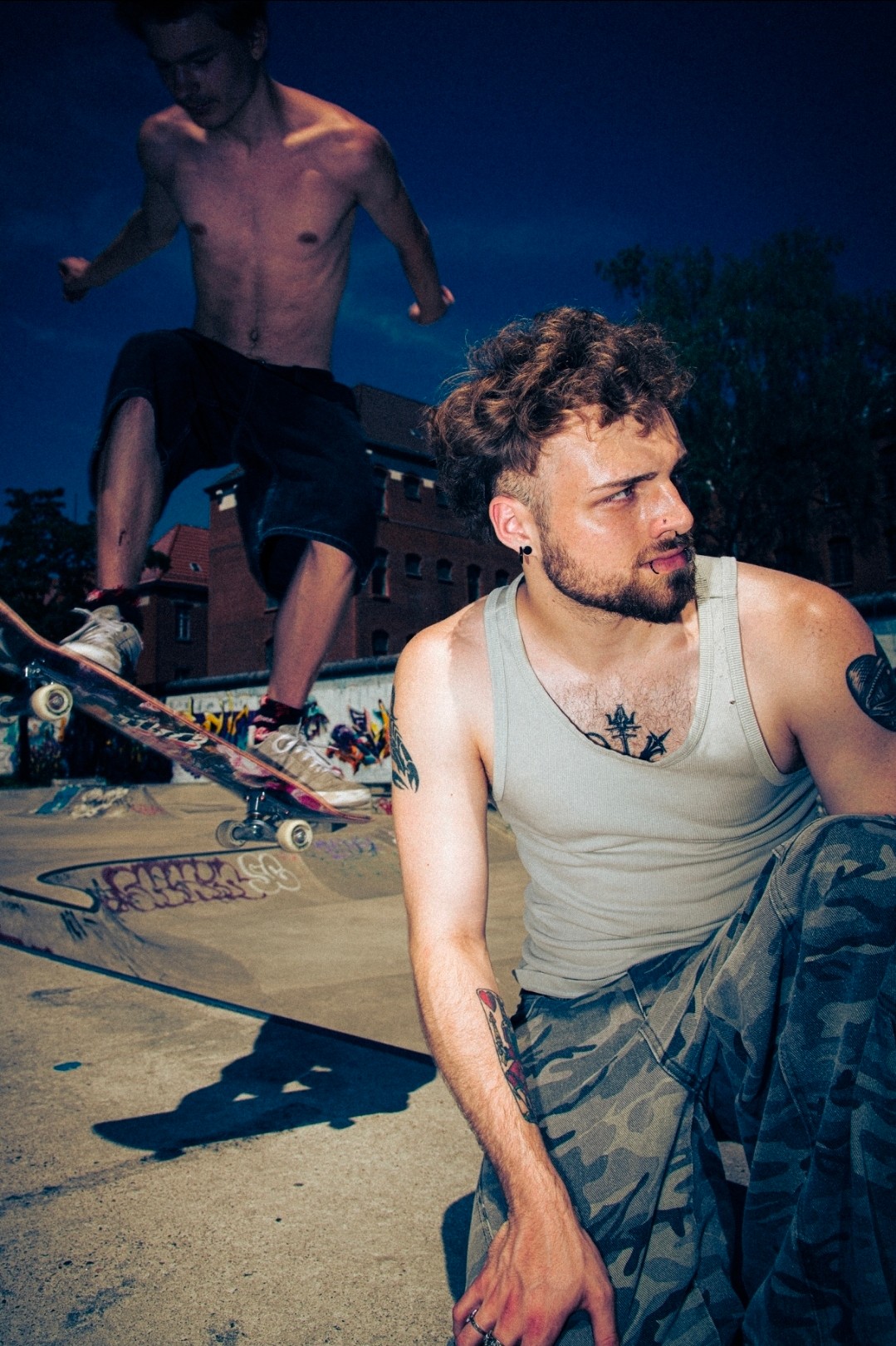 Two young men skateboarding outdoors at night, one sitting and the other standing on a skateboard, with buildings and trees in background.