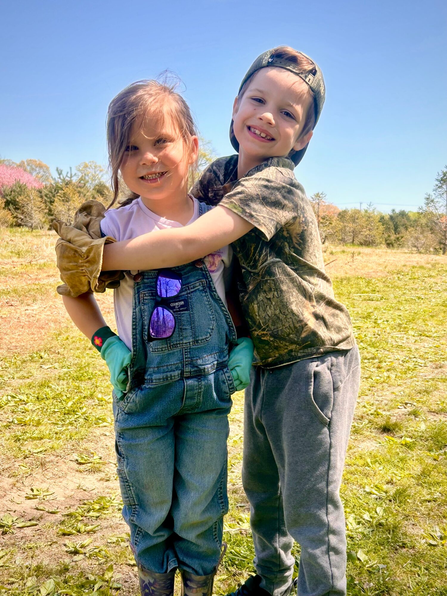 Two children hugging outdoors on a sunny day, smiling, with trees and grass in the background.