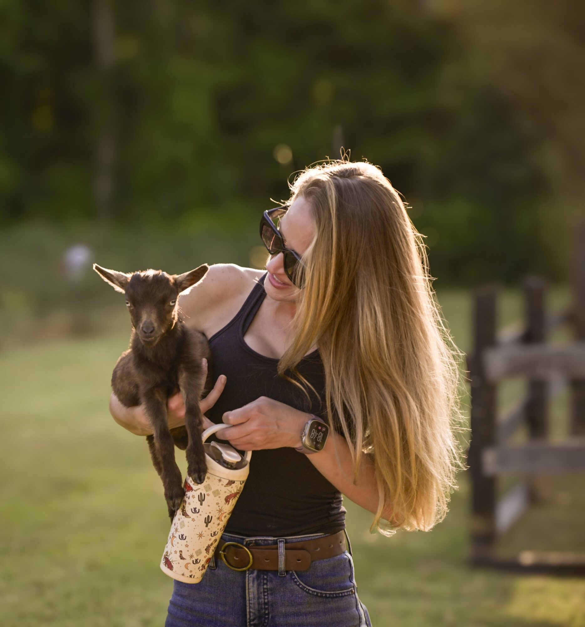 Woman holding a small goat outdoors, smiling, with a blurred natural background.