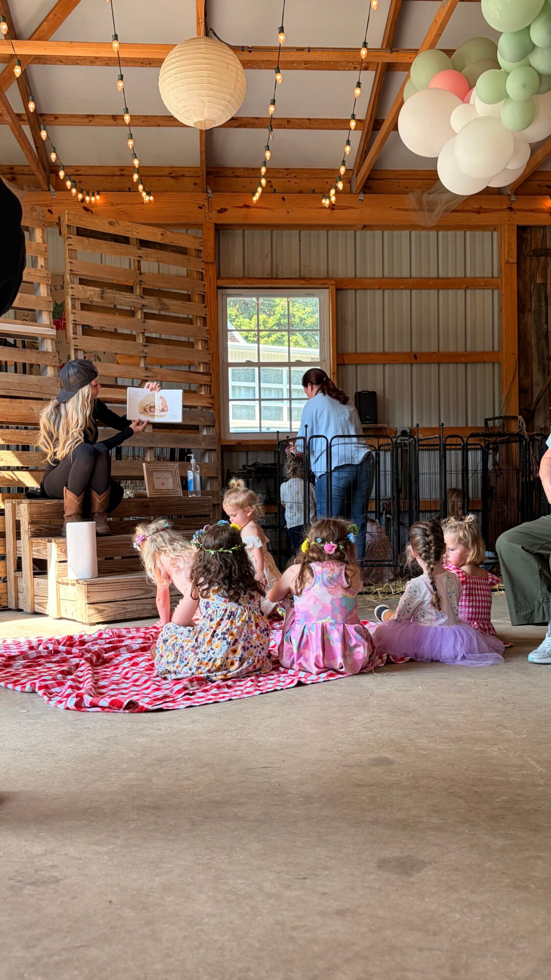 Children sitting on a pink blanket watching a person perform in a rustic wooden room with hanging paper lanterns and balloons.