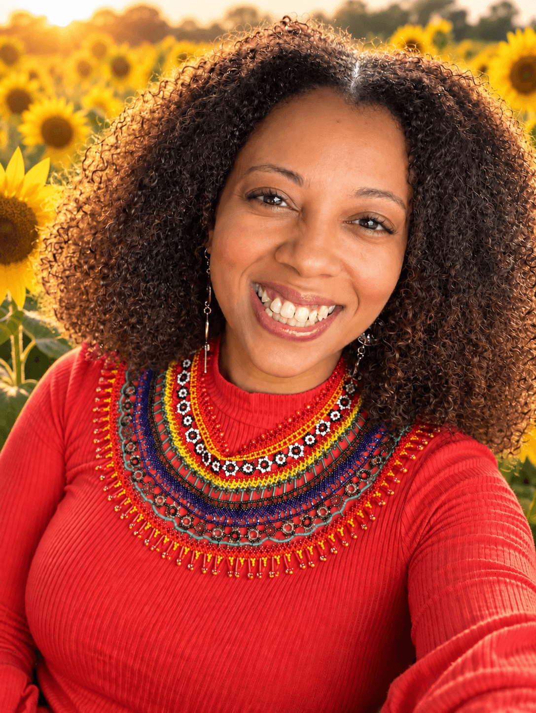 Smiling woman with curly hair in a red top, sunflower field background, bright sunlight, colorful necklace, cheerful expression