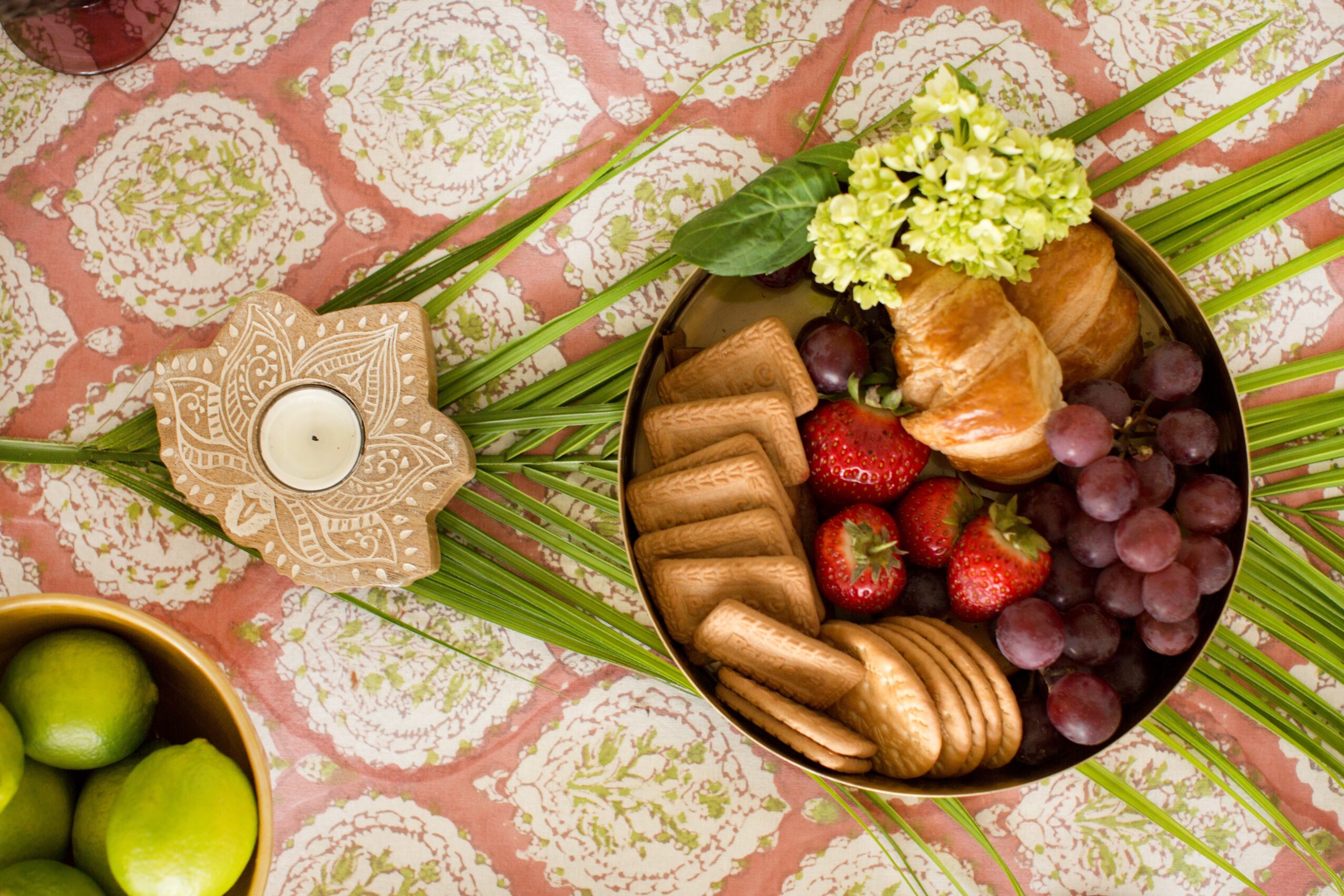 A bowl of assorted fruits and cookies on a decorative tablecloth with green leaves and a candle holder nearby.