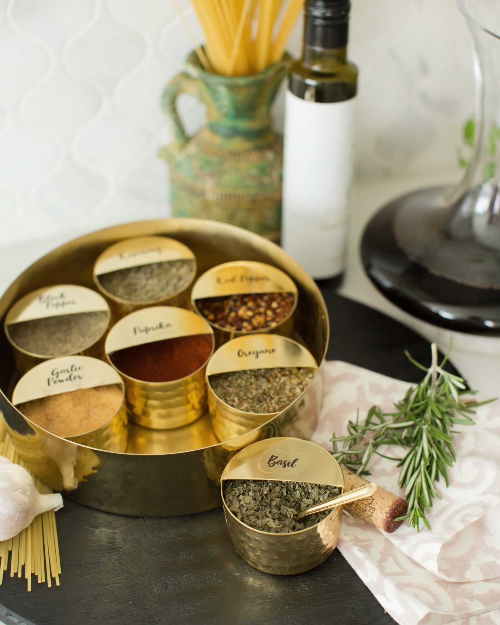 Round tins of spices arranged in a gold container, with a sprig of rosemary and kitchen items in the background.