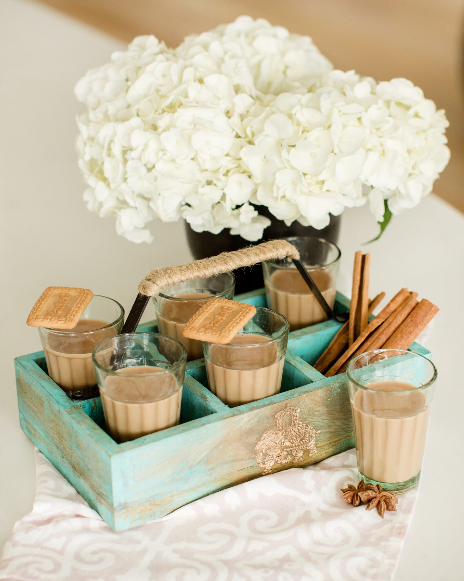 A bouquet of white flowers in a black vase, with glasses of a creamy beverage, cookies, and cinnamon sticks on a tray.