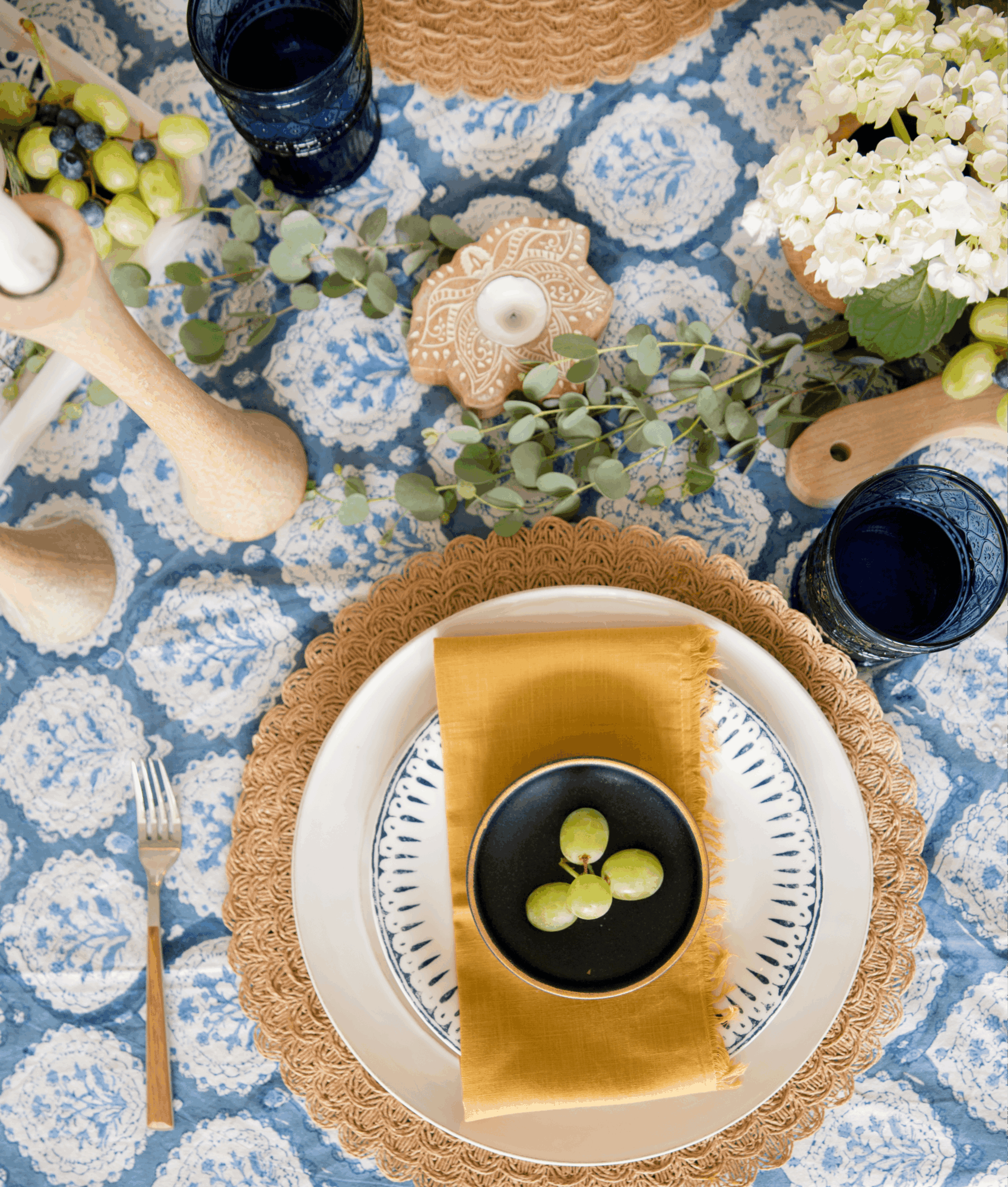 Table setting with a plate, black bowl with grapes, gold napkin, and floral decorations on a blue patterned tablecloth.