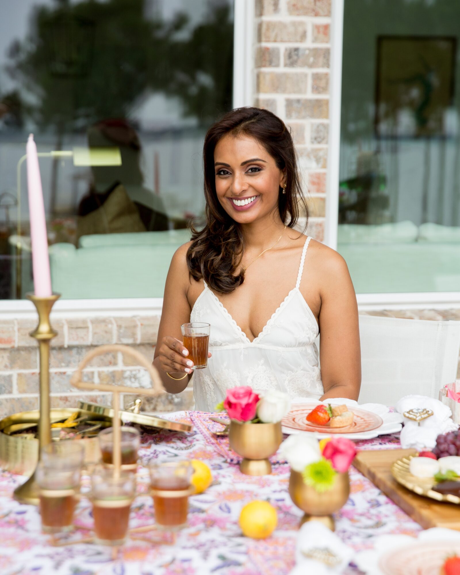 Smiling woman in white dress holding a drink at a table with food and flowers outside.
