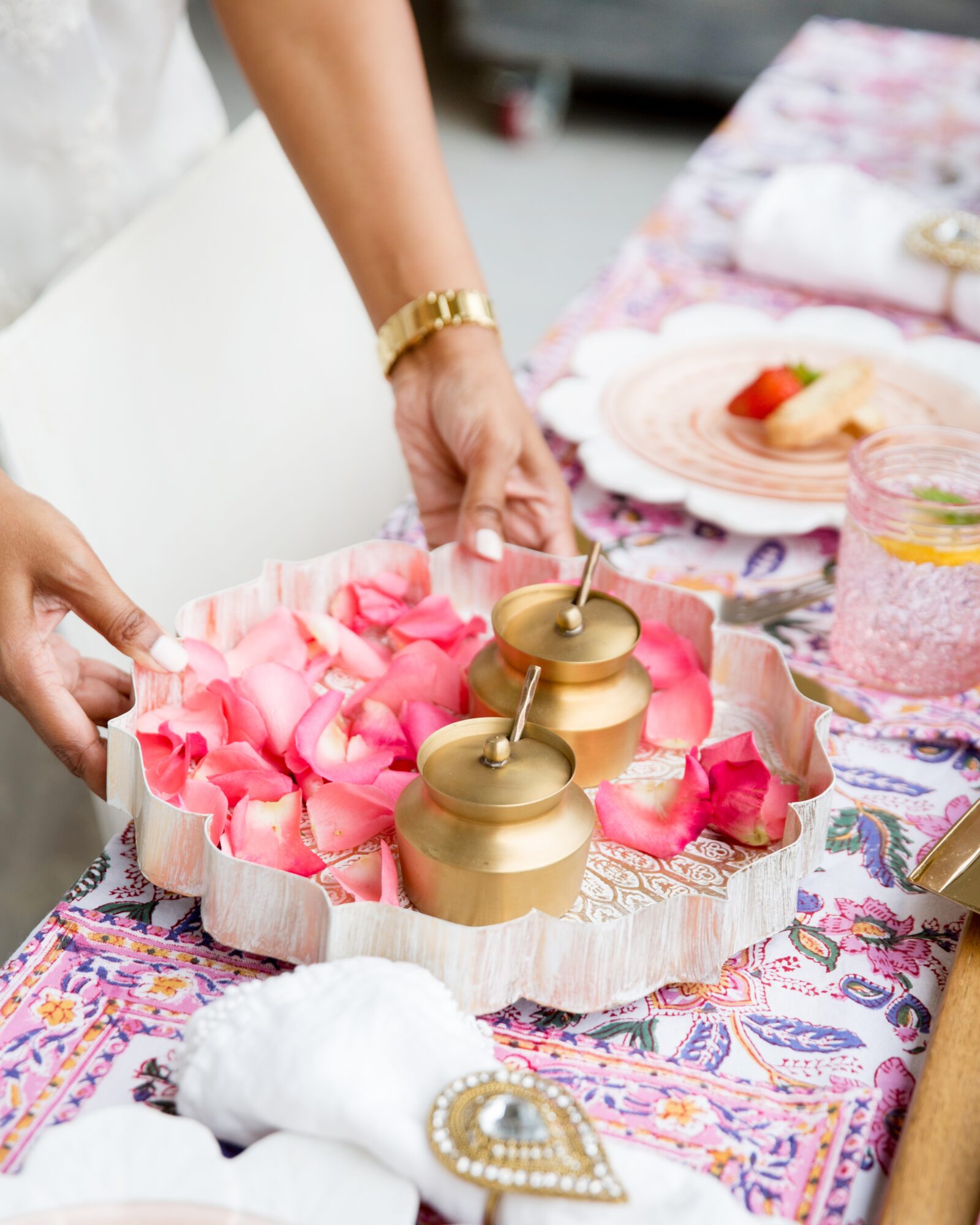 Person arranging pink rose petals around two gold candle holders on a decorated table, with plates and glasses visible.