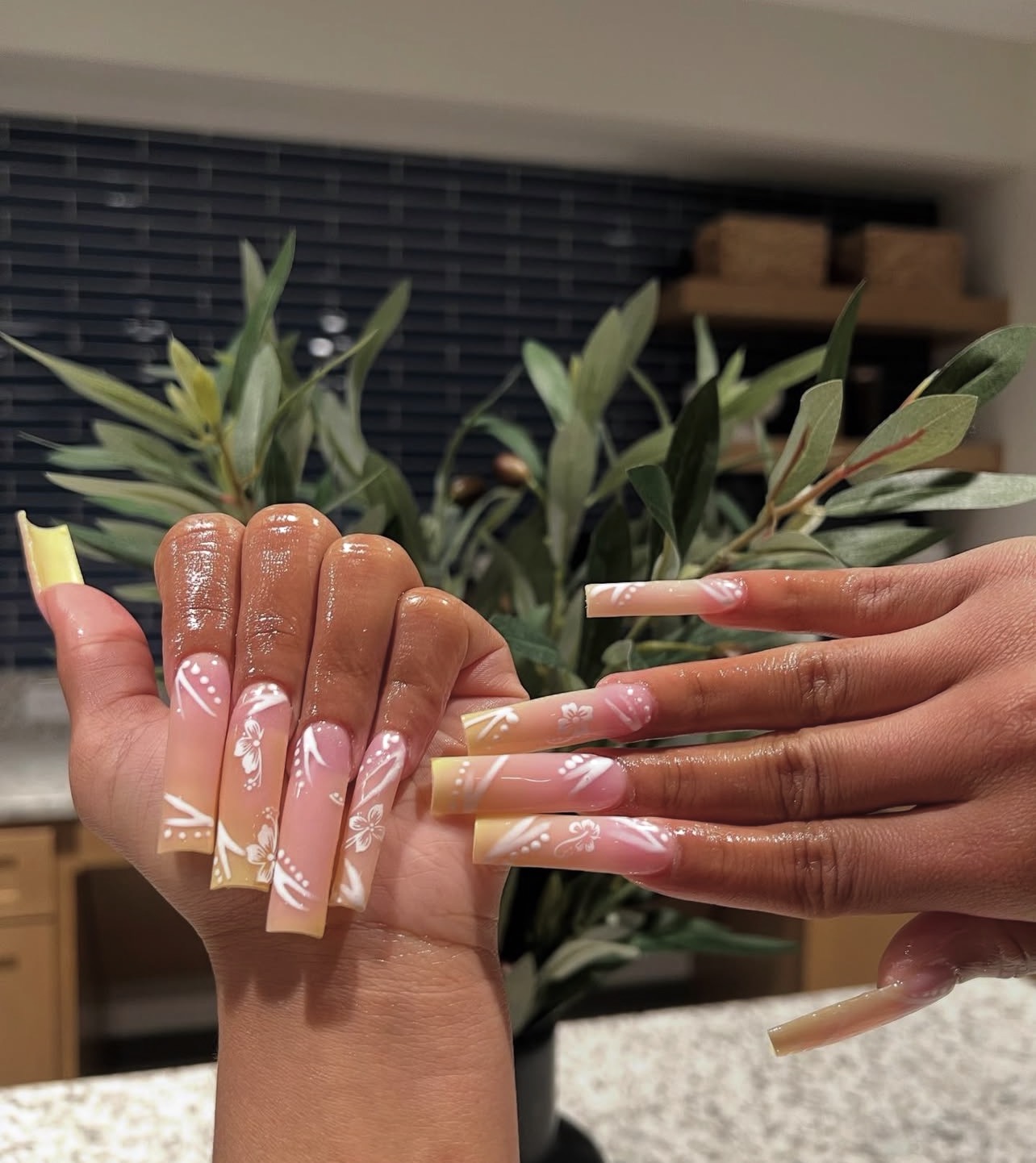 Two hands with long, decorated nails, one with a pink and white design, the other with a natural look, in front of green plants.