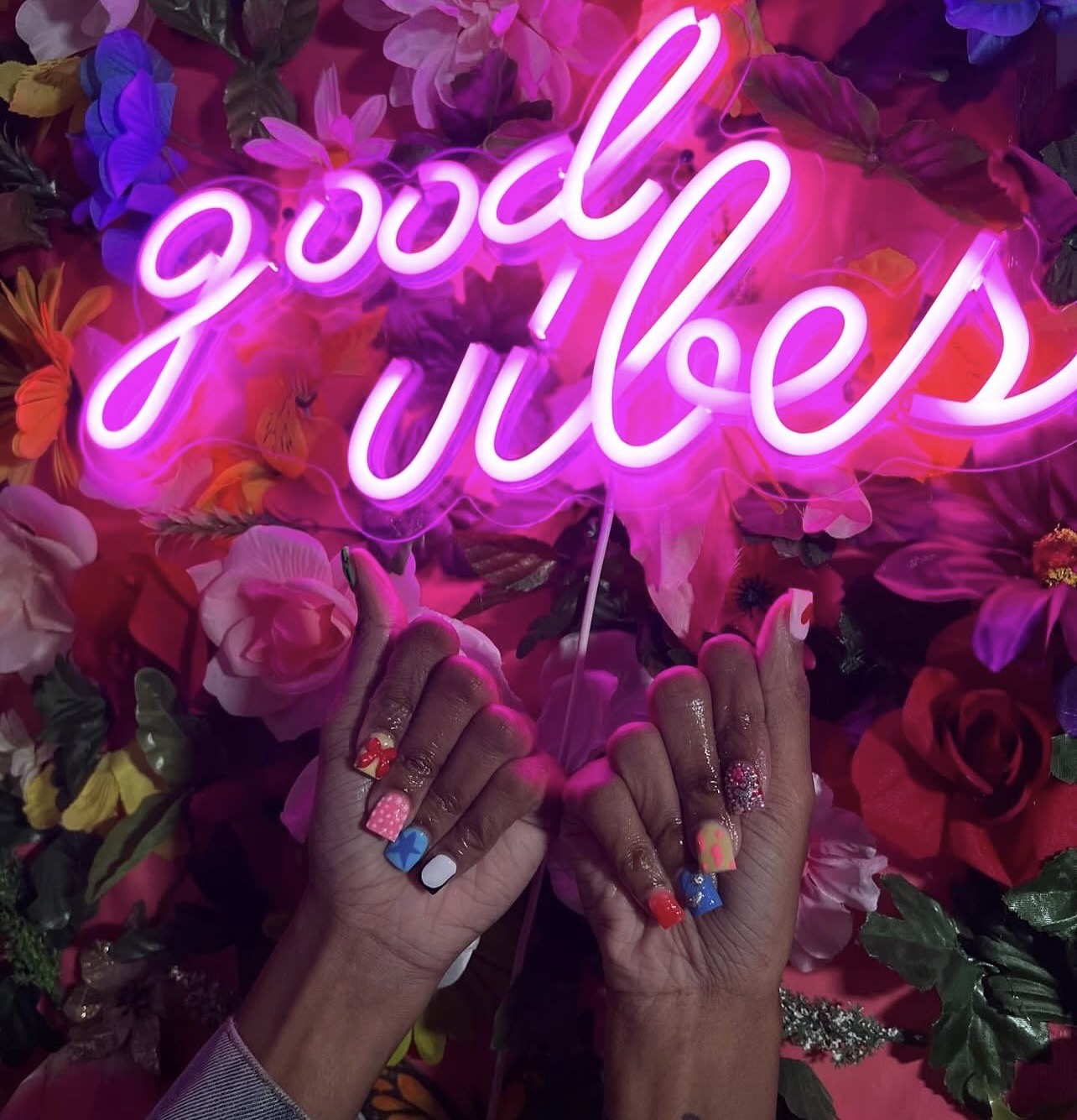Two hands with colorful nail polish hold a pink neon sign that says 'good vibes' against a background of flowers.