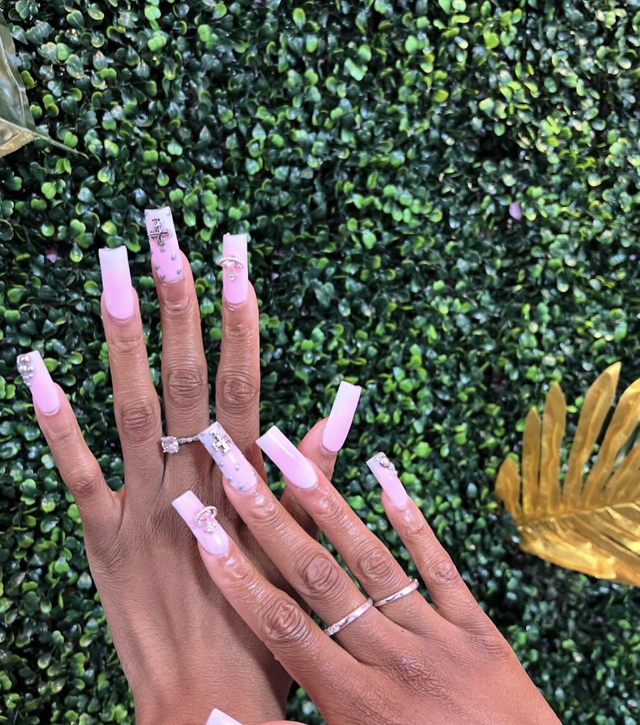 Hand with long, pink, decorated nails and rings, against a green leafy background with a yellow decorative element on the right.