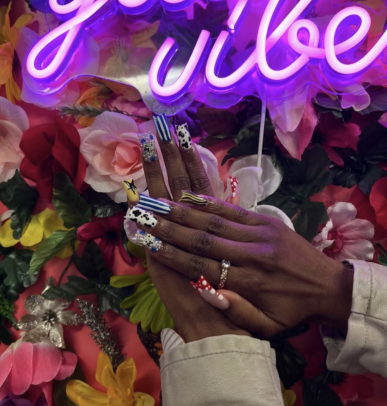 Person's hand with decorated nails and ring, touching a floral background with a neon sign, flowers, and a cake.