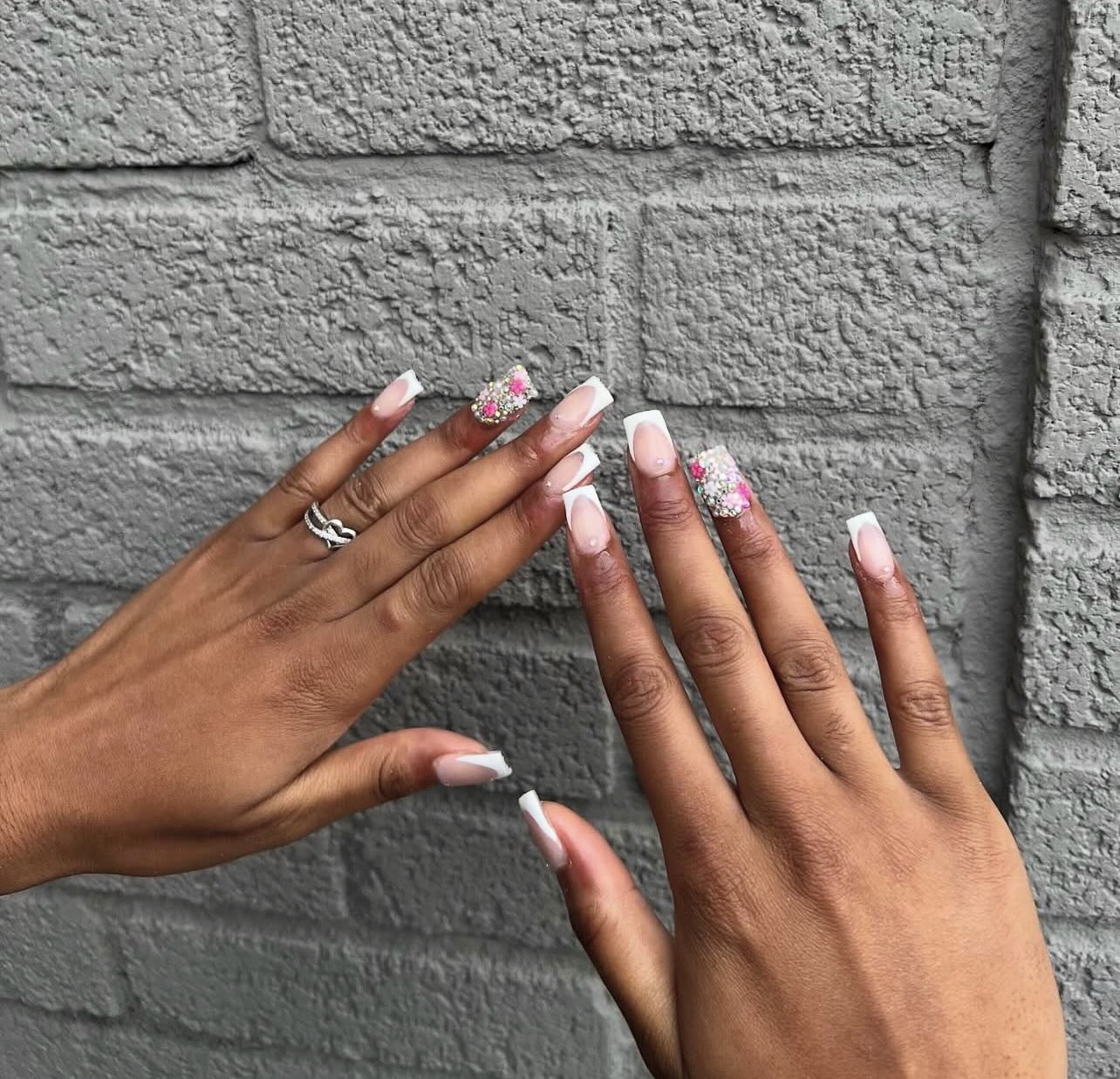 Two hands with decorated nails and rings, showing nails with pink and white floral designs, against a gray brick wall.