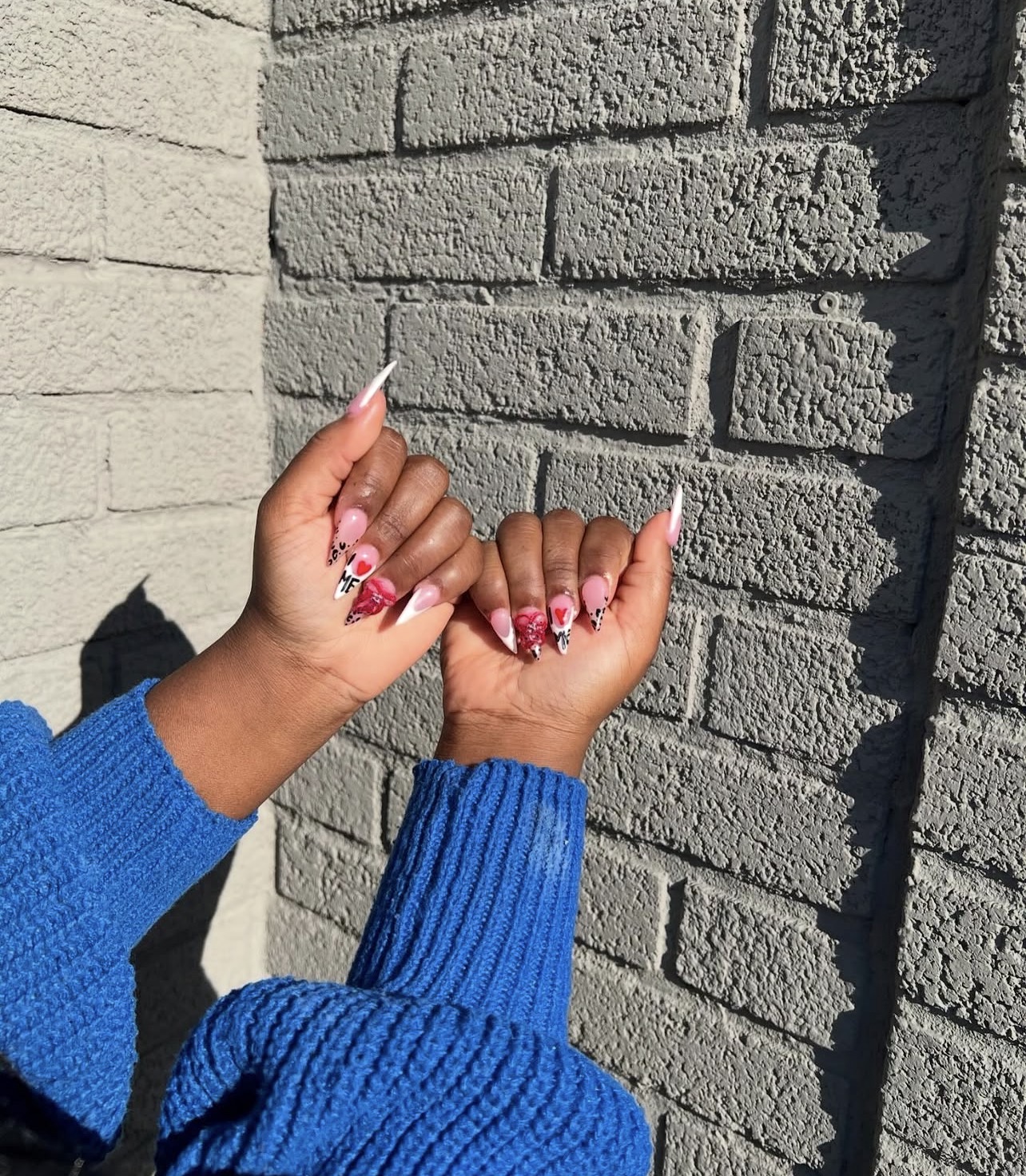 Two hands with pink and white nail art, wearing a blue sweater, are raised against a brick wall background.