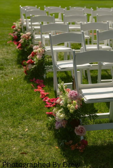 White chairs decorated with pink and white flowers arranged along a grassy outdoor setting, likely for an event.