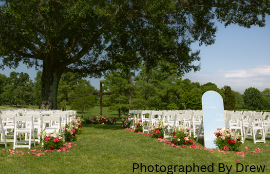 Outdoor wedding setup with white chairs, flower arrangements, and a large tree in the background.