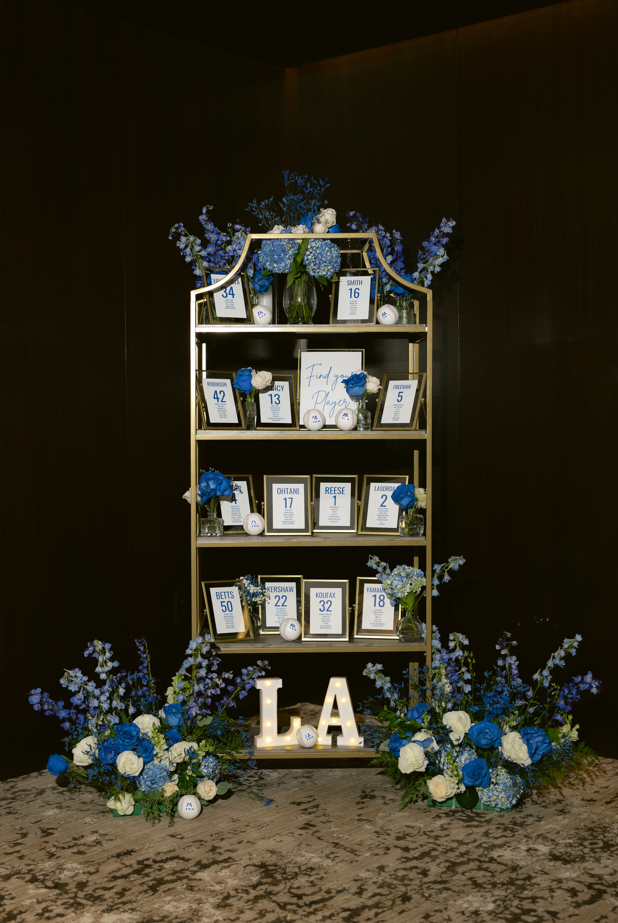 Decorative display with shelves holding framed photos and blue flowers, surrounded by floral arrangements, and illuminated letters spelling 'LA'.