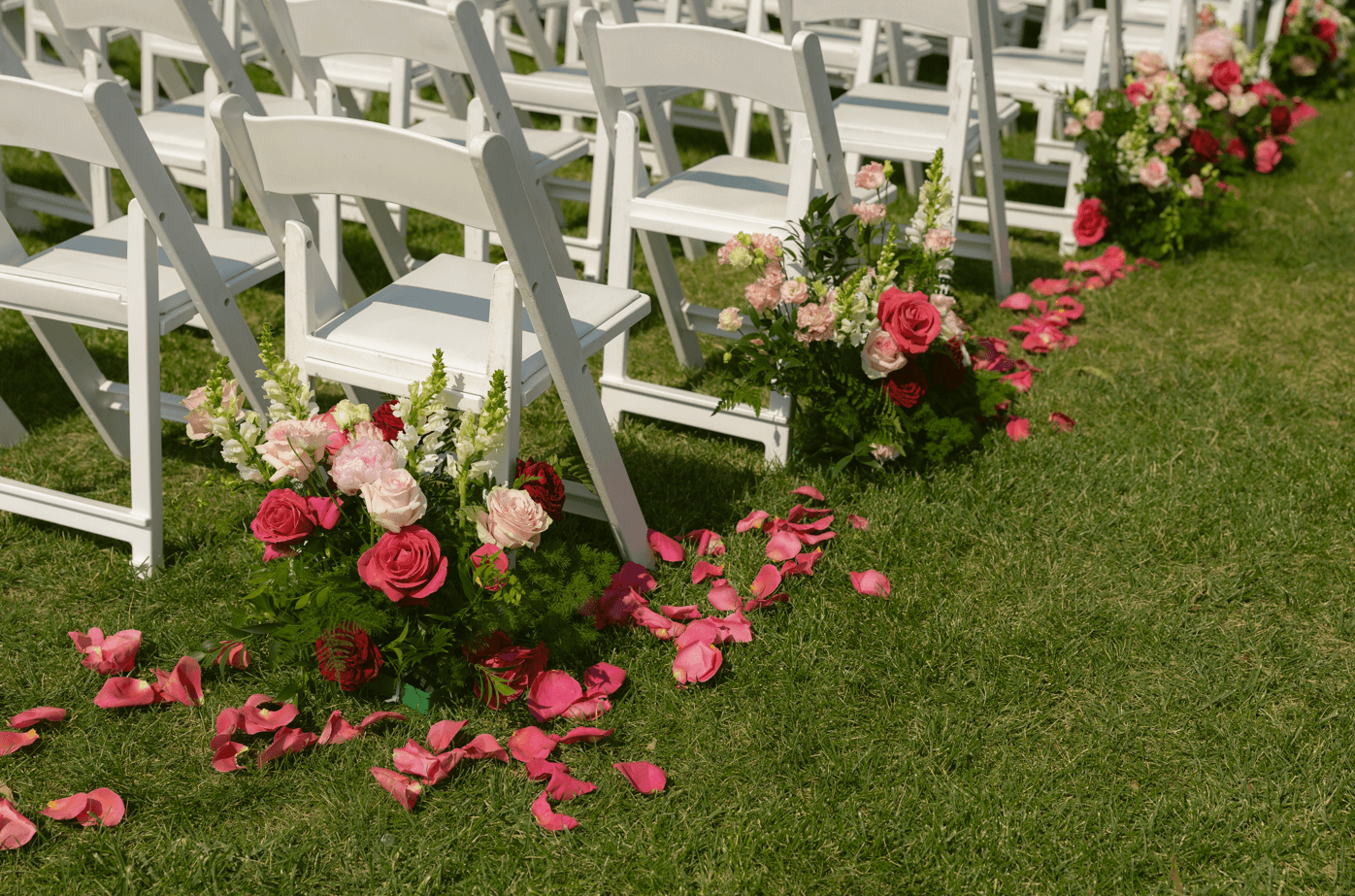 White chairs with floral arrangements and pink rose petals on grass at an outdoor event.
