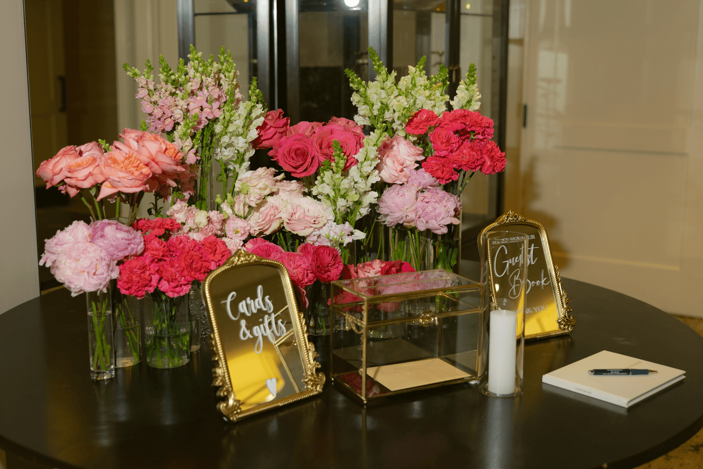 Table with pink and white flowers in glass vases, two gold frames with signs, and a small box, in a room with glass doors