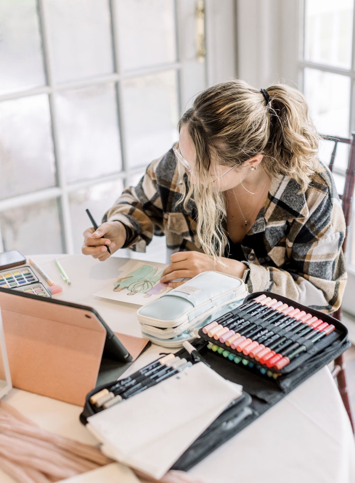 Person with long hair in a plaid shirt working at a desk with art supplies and a laptop, near large windows.