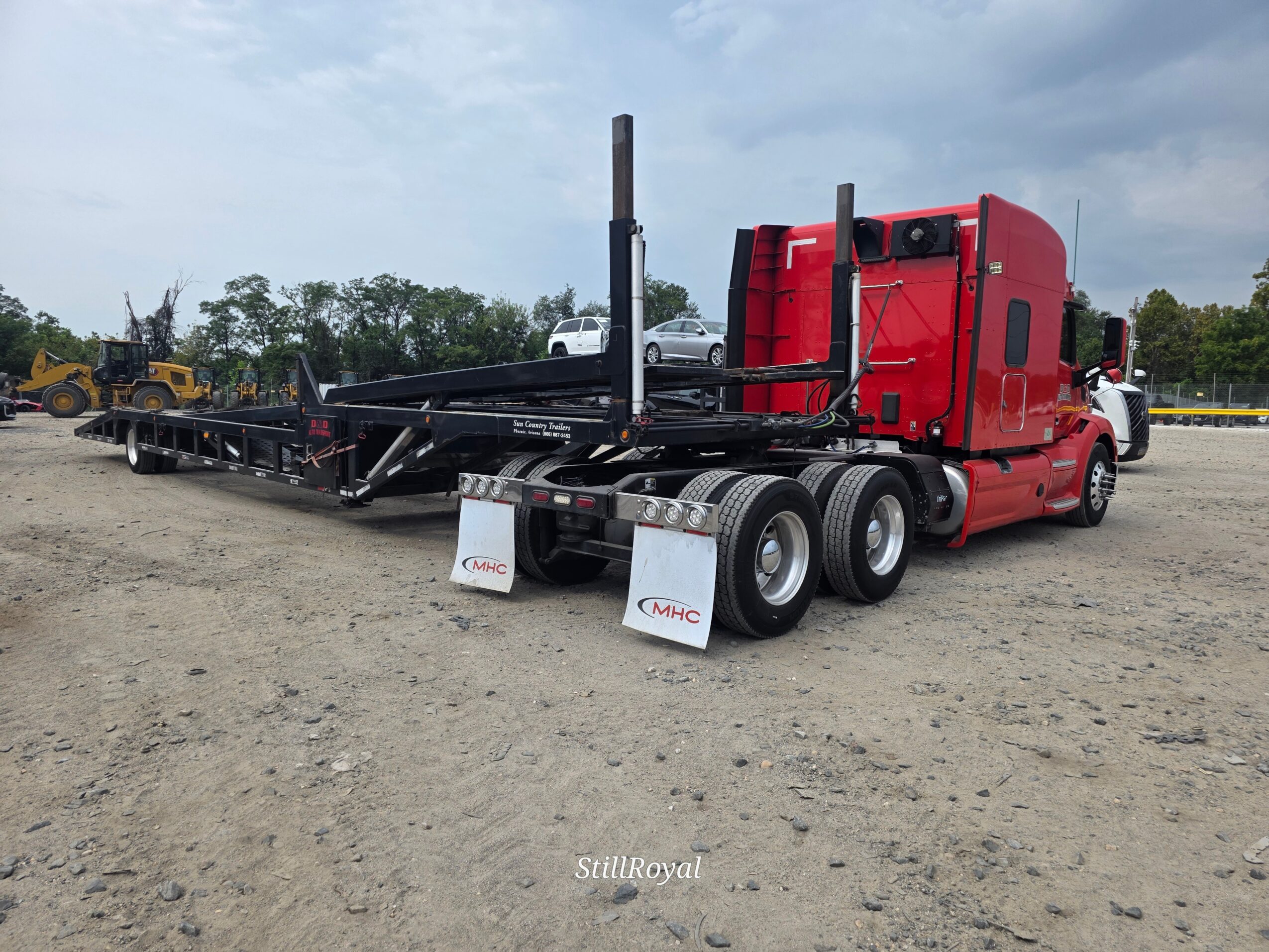 Red semi-truck with flatbed trailer parked on dirt lot, cloudy sky background, with trees and construction equipment nearby.
