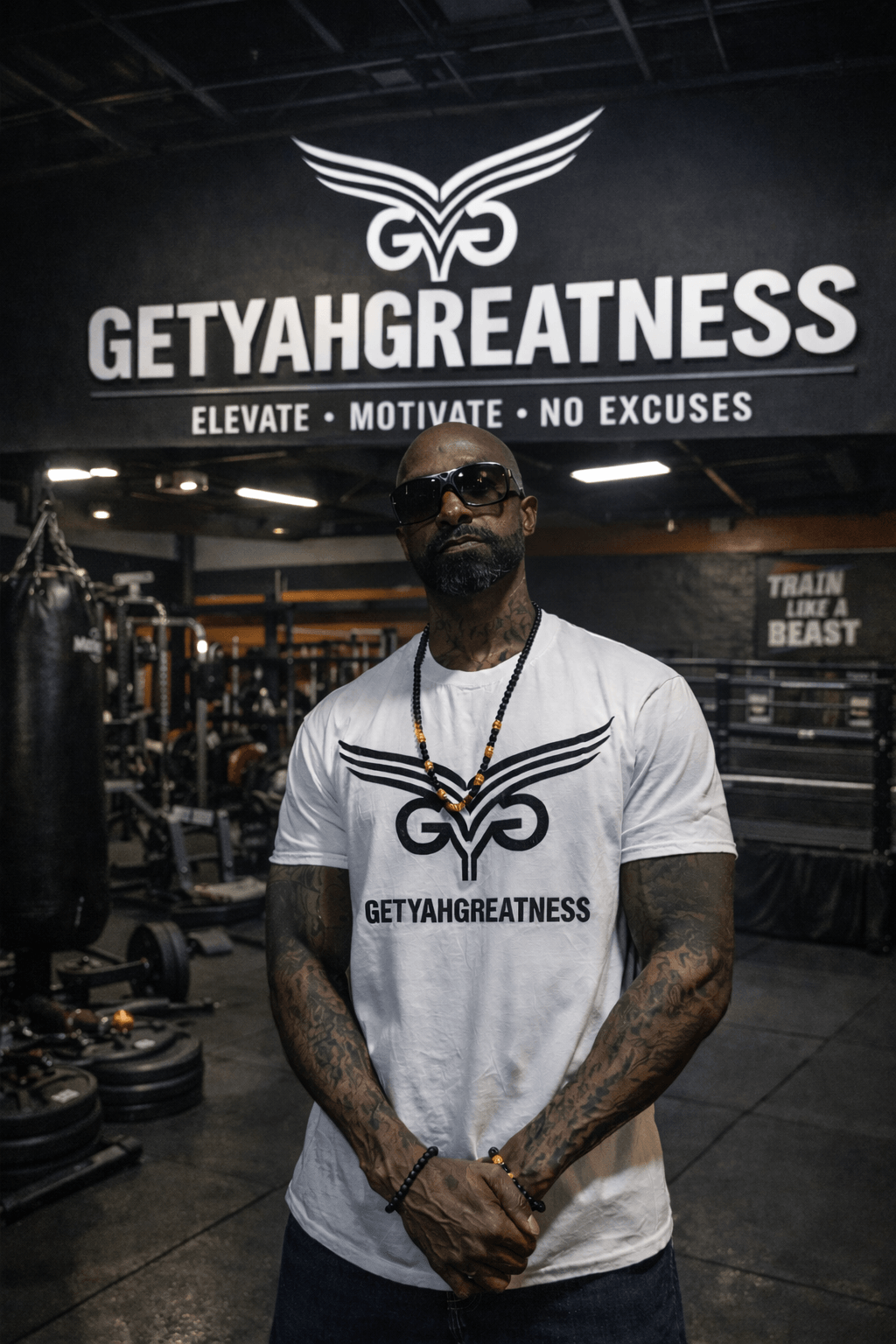 Man with sunglasses and tattoos standing in gym, large sign reads 'GETYANGREATNESS' and motivational phrases behind him.