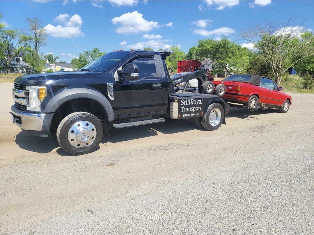 Black flatbed truck with a red car attached, parked on a dirt road with trees and blue sky in background.