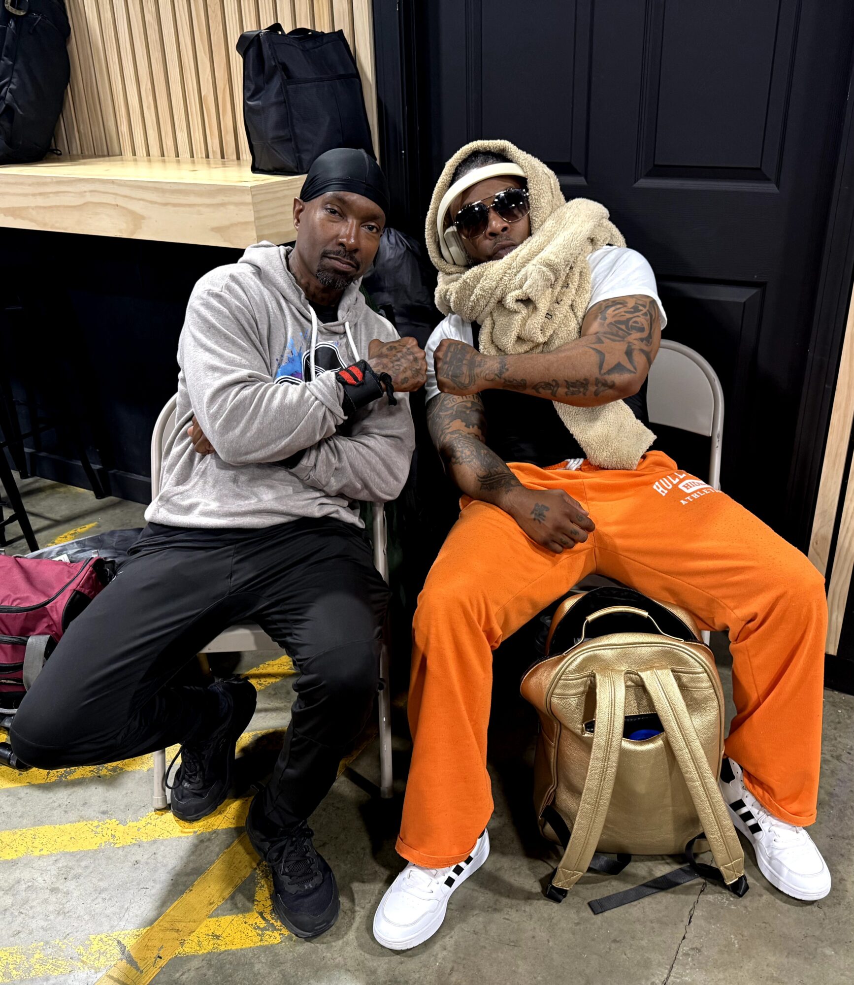 Two men sitting on chairs, one wearing a gray hoodie and black pants, the other in orange pants, white sneakers, sunglasses, and a large scarf. They are indoors with a black door and wooden paneling behind them.