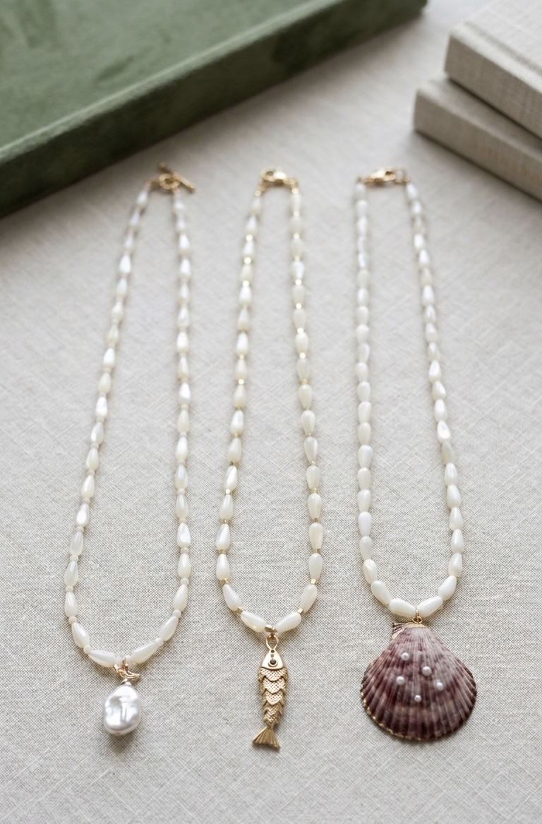 Three pearl necklaces with different pendants on a light surface, with books and a green box nearby.