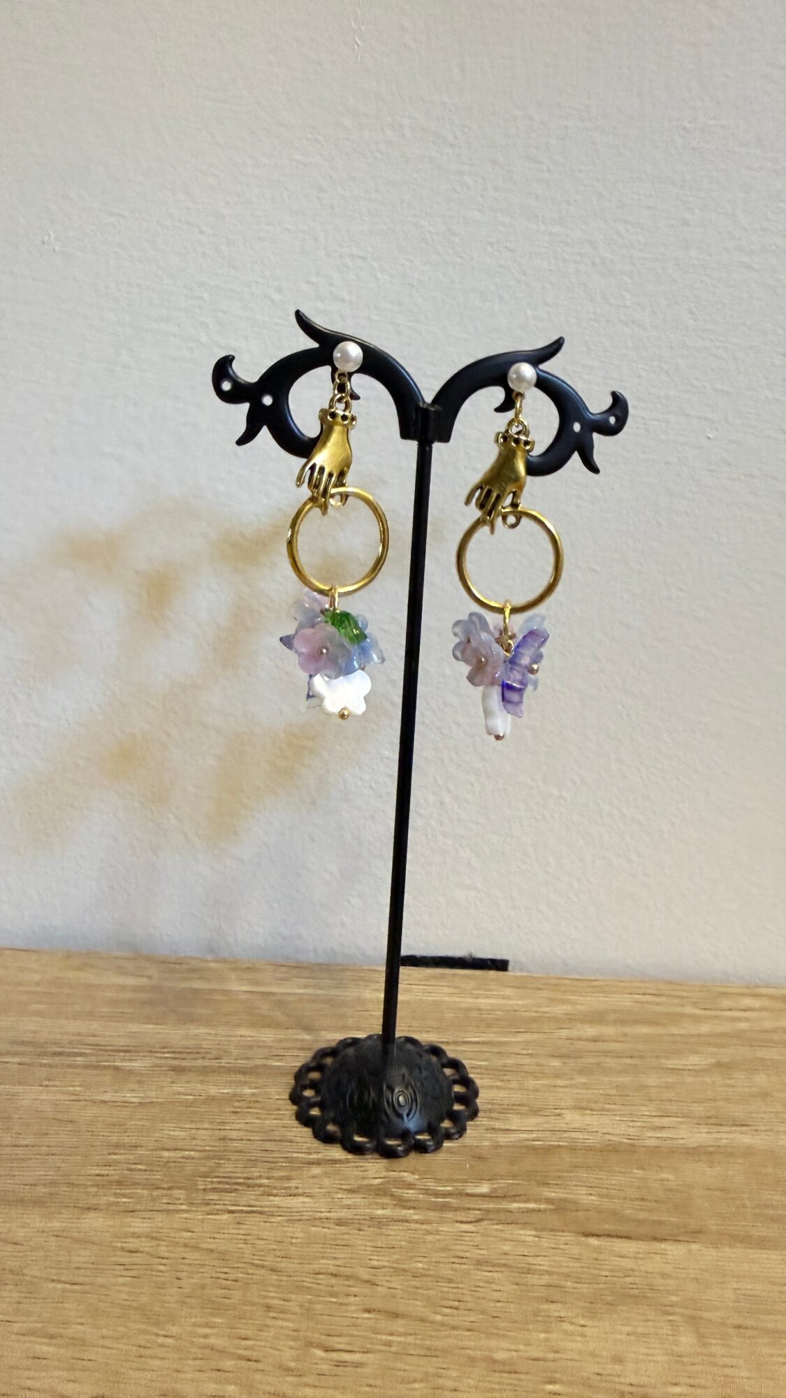 Earrings with purple and clear crystal charms hanging from gold hoops on a black display stand, on a wooden surface.