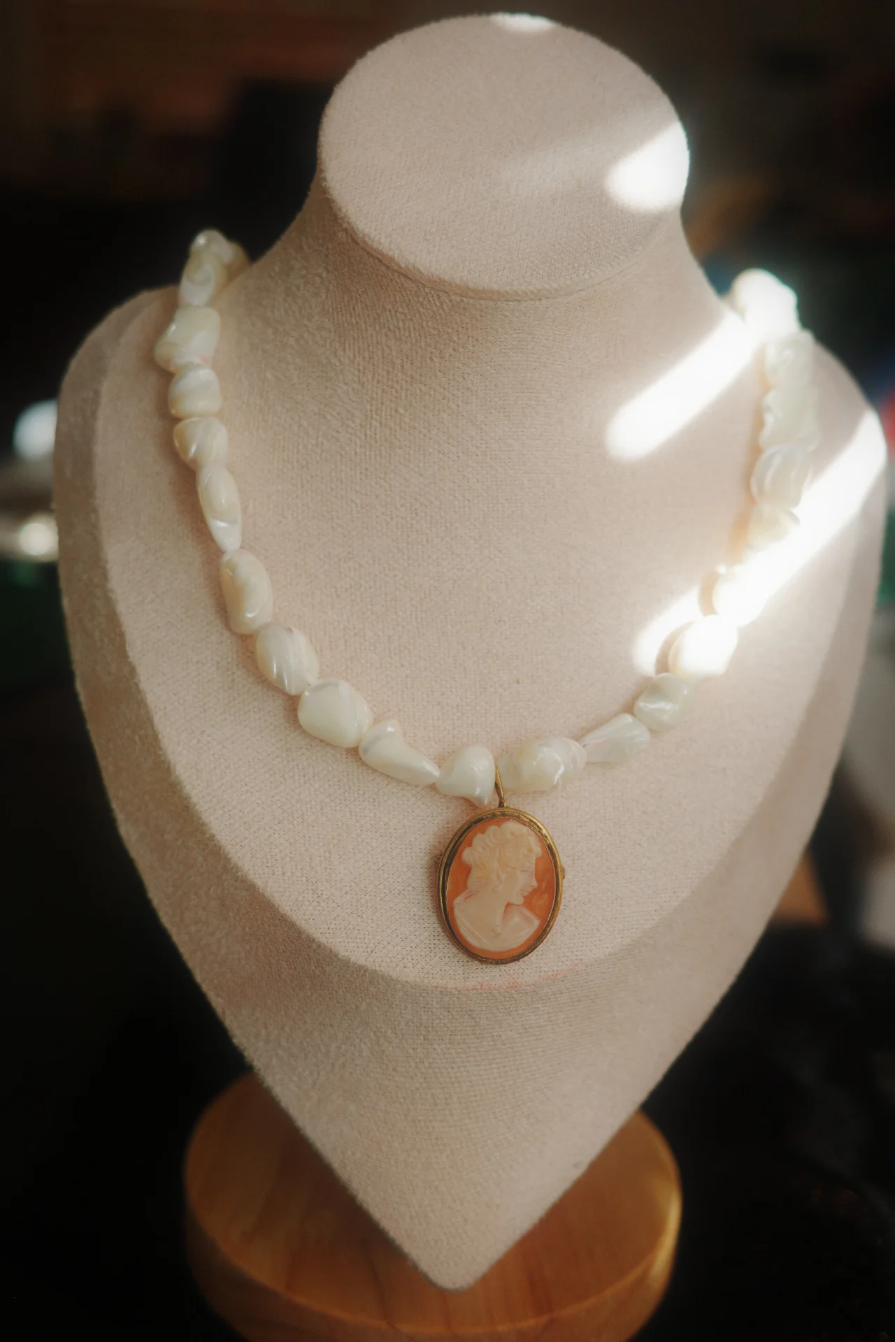 Beige necklace with a pendant displayed on a white bust, illuminated by light, with a dark background.