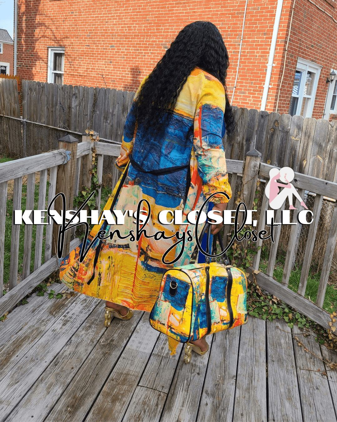 Person with long curly hair wearing a colorful coat and carrying a matching bag on a wooden deck, fence, and brick building background.