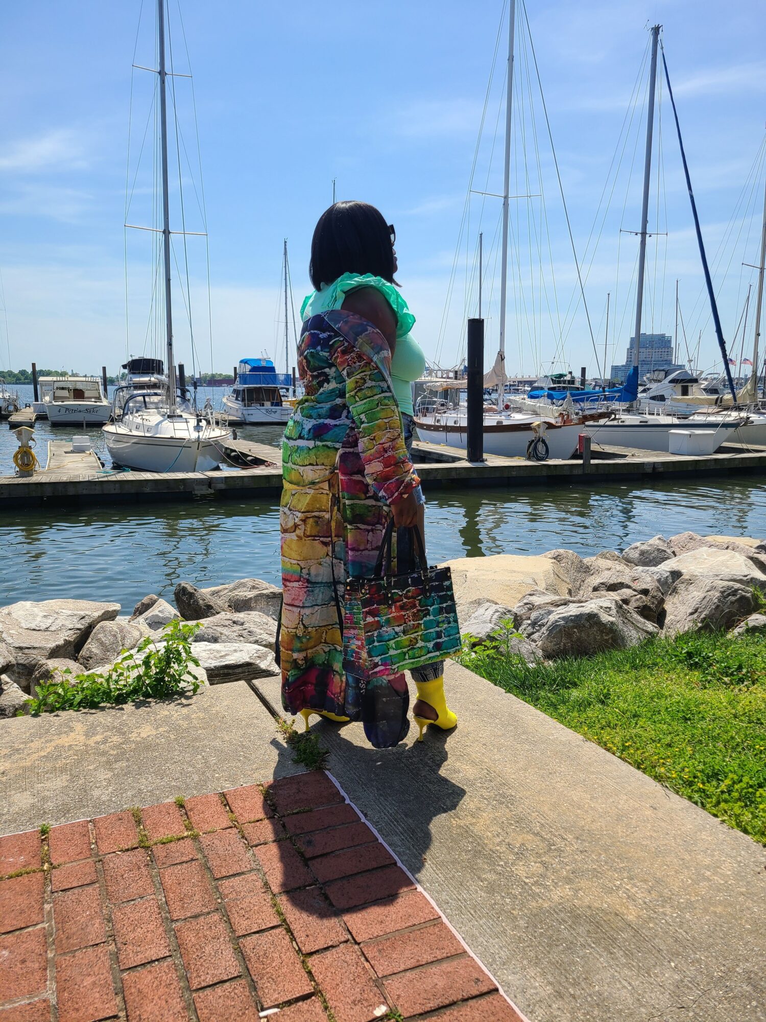 Person standing on a dock near boats and water, wearing colorful patchwork clothing, facing away from the camera.