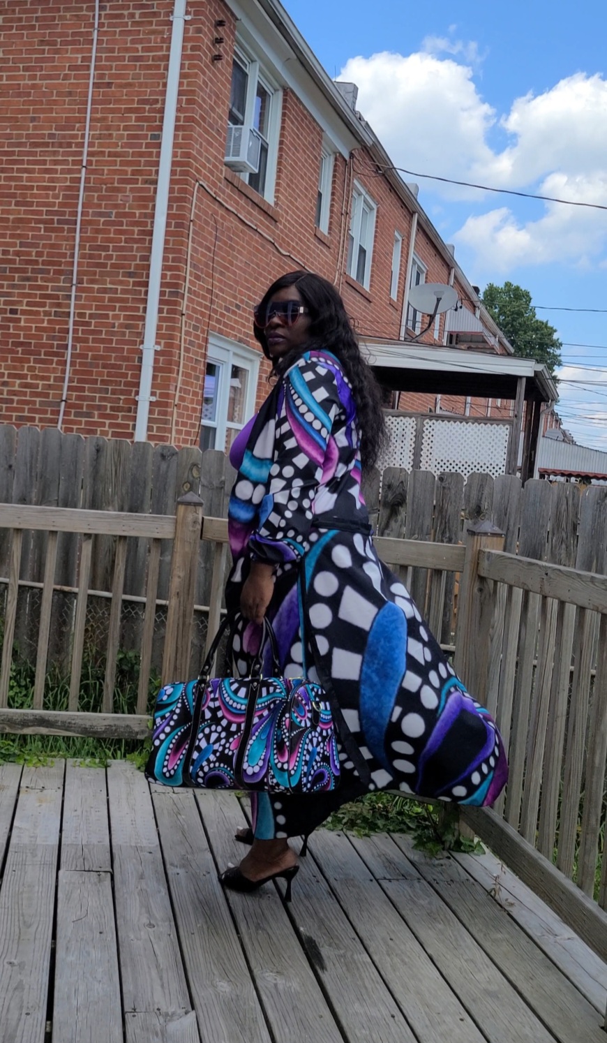 Woman with sunglasses standing on a wooden deck, holding large patterned bags, in front of a brick building.
