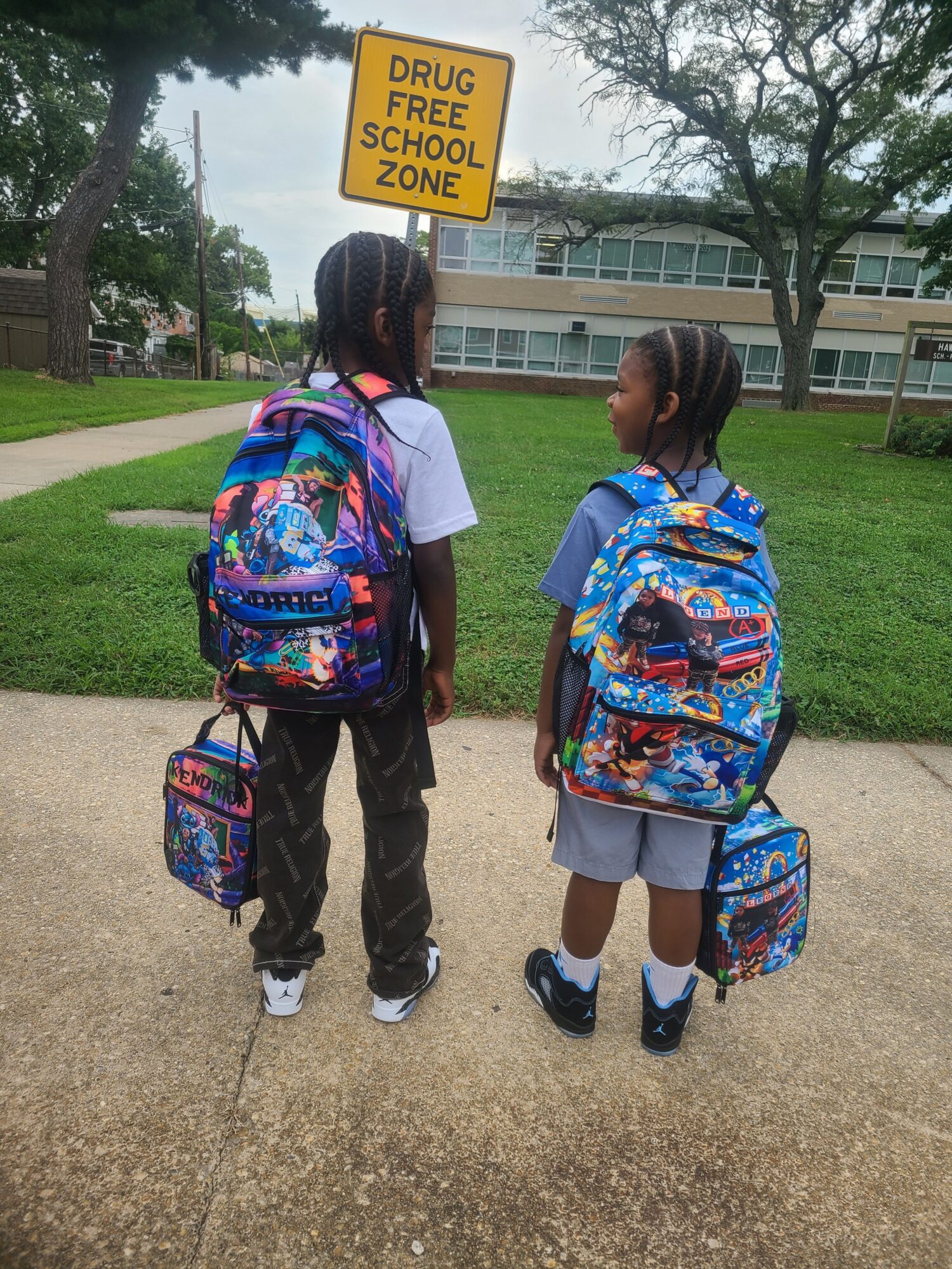 Two children with backpacks walking on a sidewalk near a school building, one holding a yellow sign, trees in background.