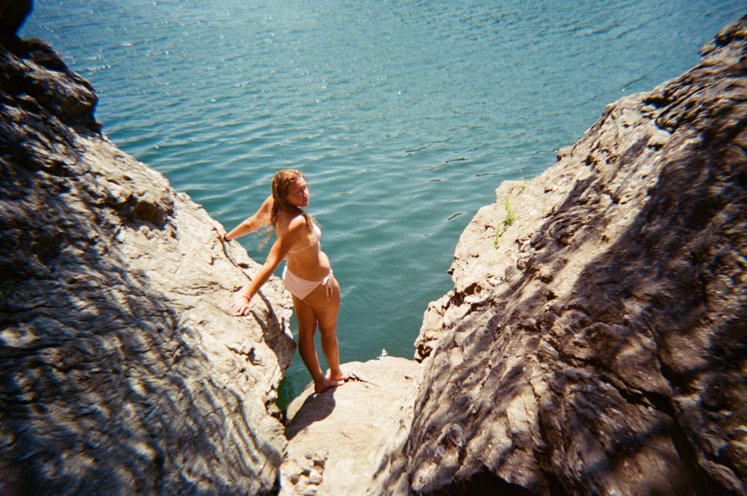 Woman in a bikini standing between rocks near water, holding onto a rock, with water in the background.