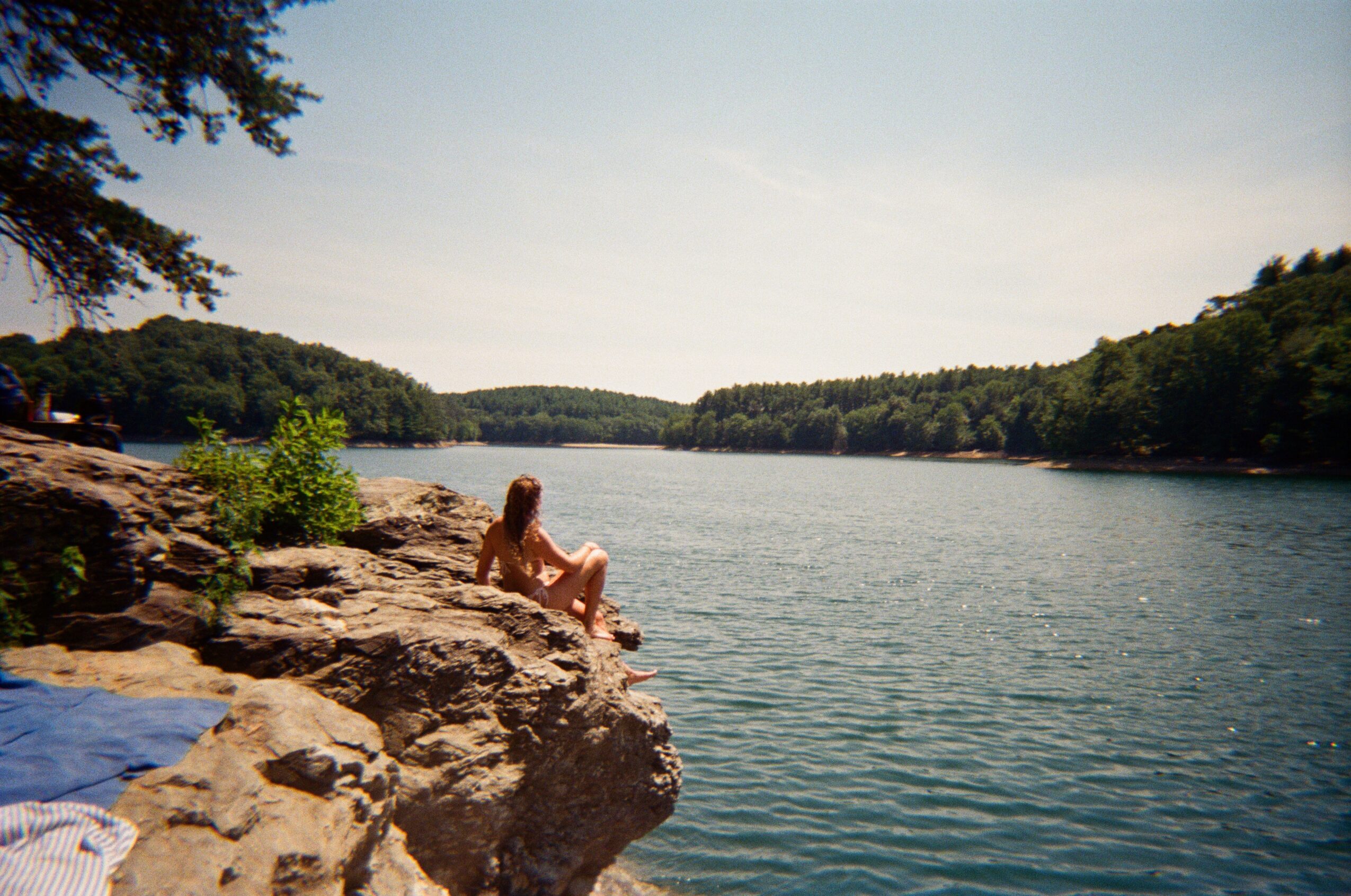 Person sitting on rocks by a river with trees and hills in the background.