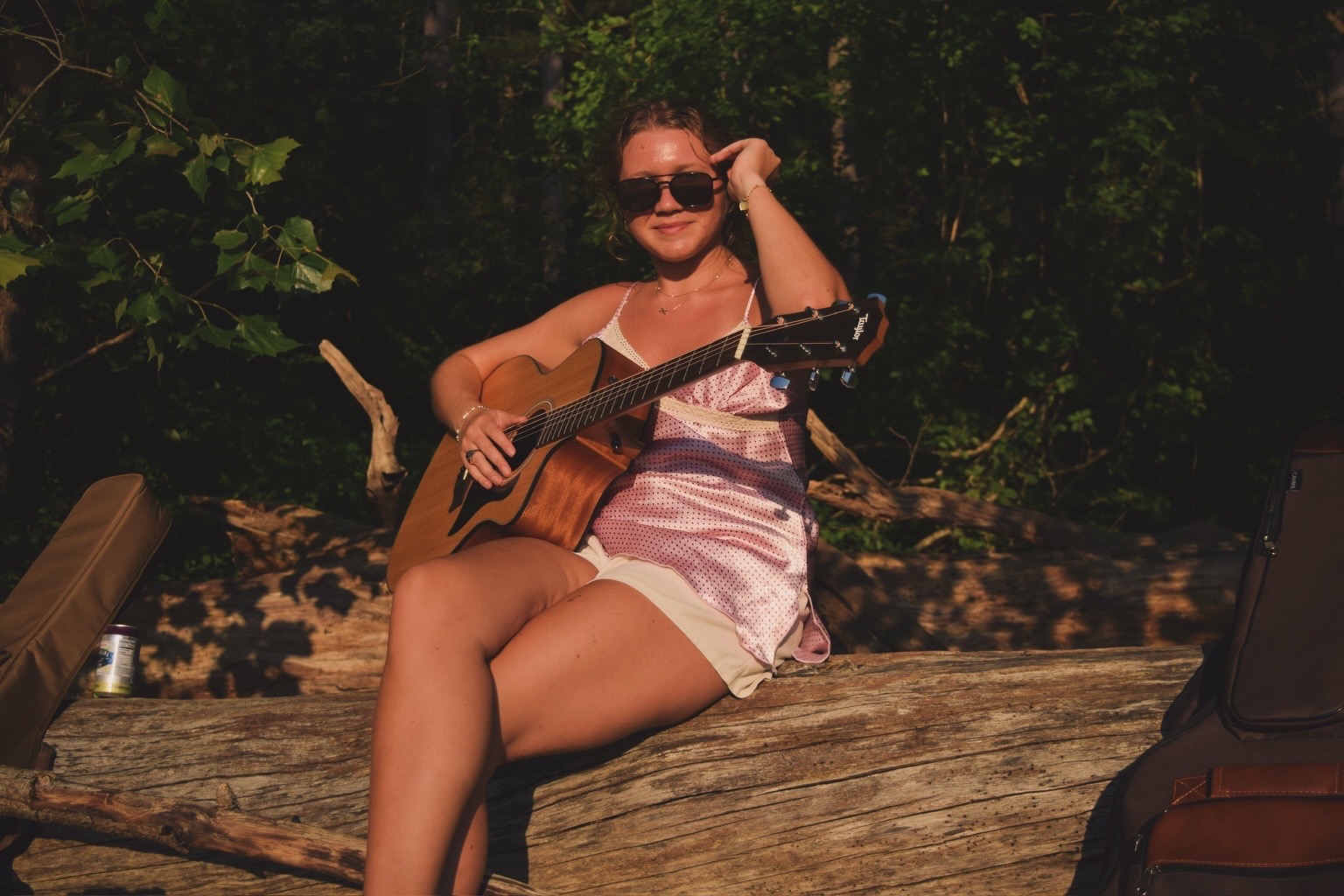 Woman sitting on a log outdoors, playing guitar, wearing sunglasses and a light-colored dress, surrounded by greenery.