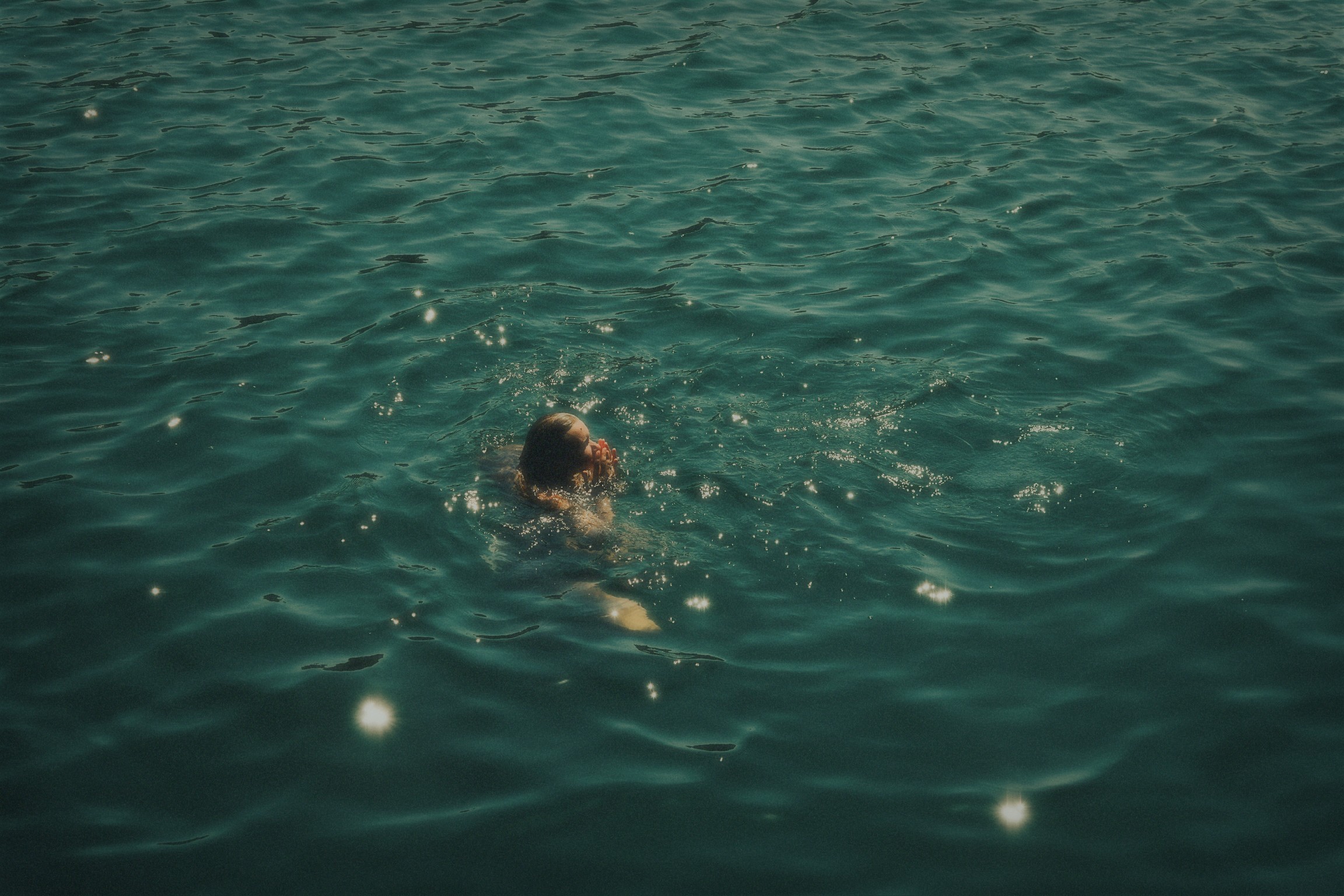 Person swimming in a body of water with ripples and reflections, facing downward with head submerged.