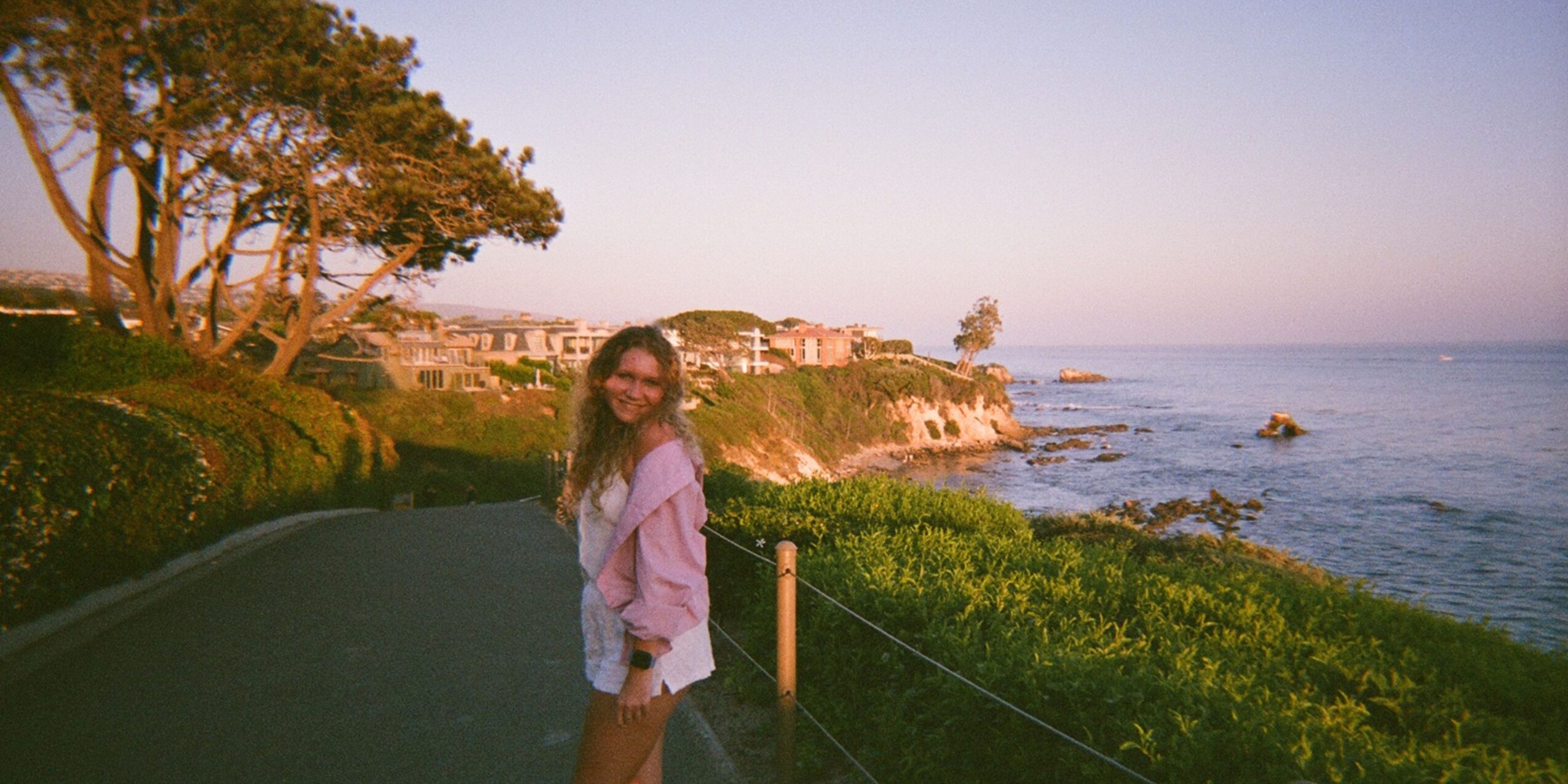 A woman stands on a coastal path with trees and buildings in the background, overlooking the ocean during sunset.